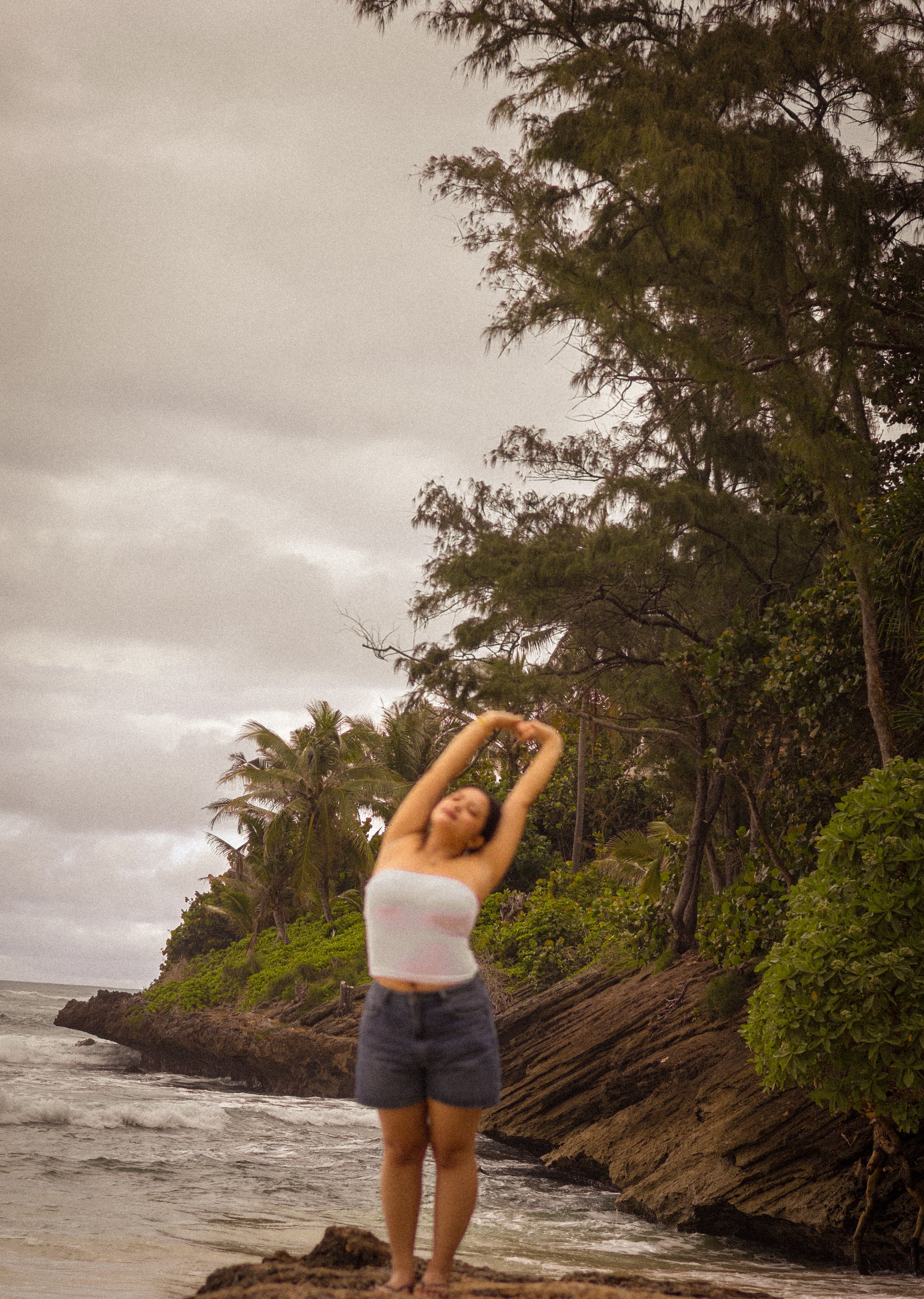 A woman in a white strapless top and denim shorts stretching her arms above her head on a beach with rocky shoreline, lush green trees, and cloudy sky.