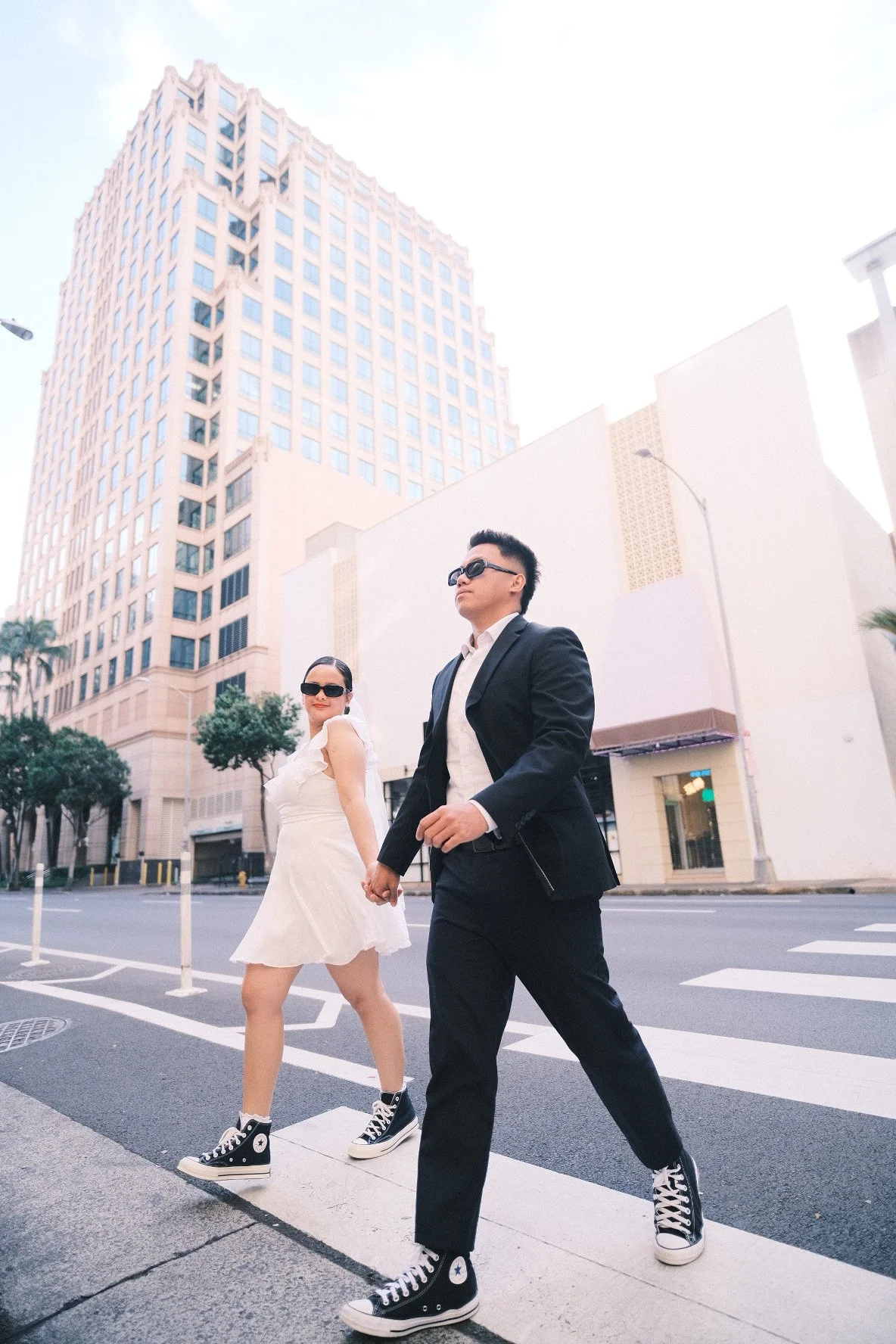 A stylish man and woman walking across a city crosswalk, holding hands, with tall buildings and a bright sky in the background.