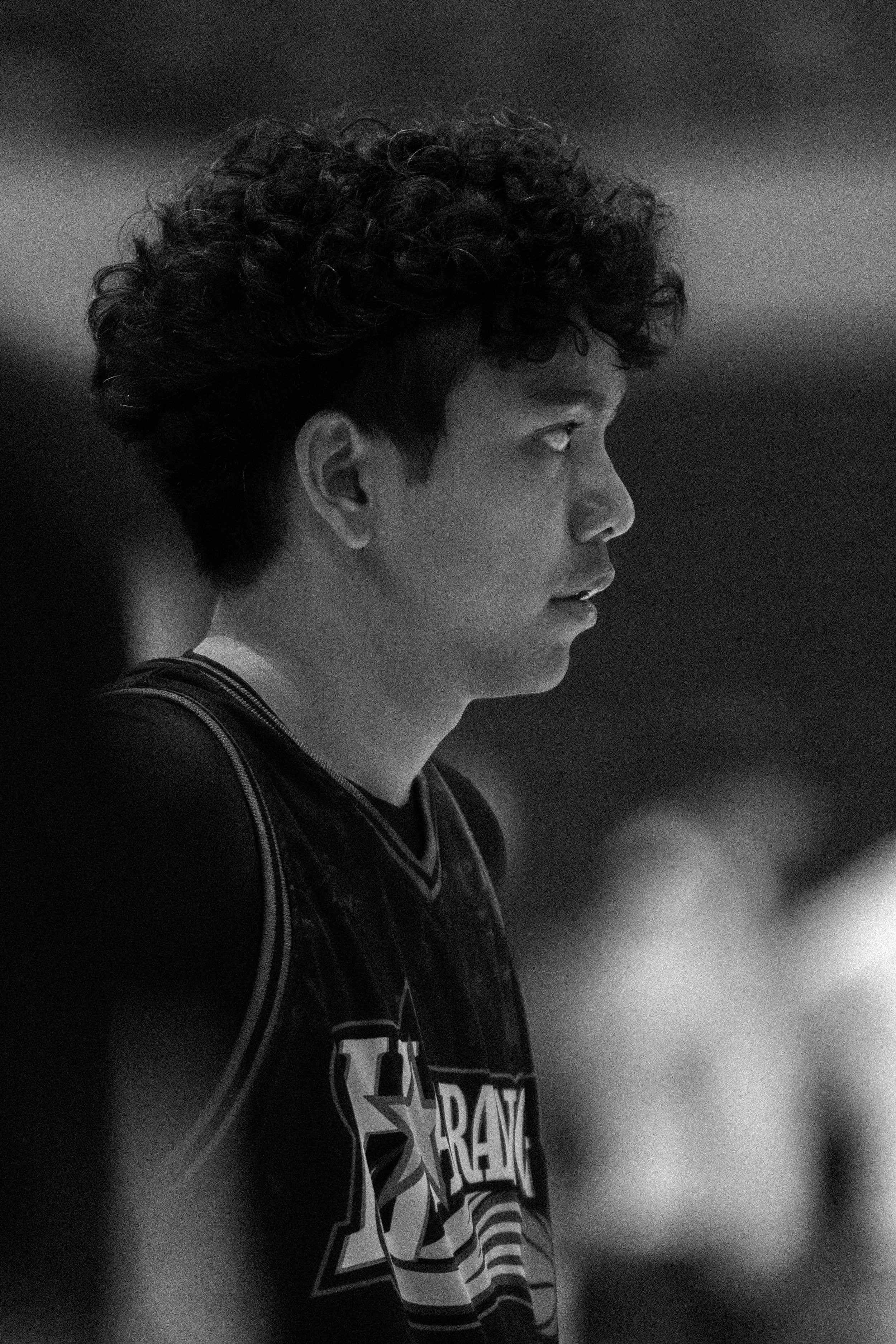 A young man with curly hair looking to the right, wearing a sports jersey with the logo 'UCLA' in black and white.