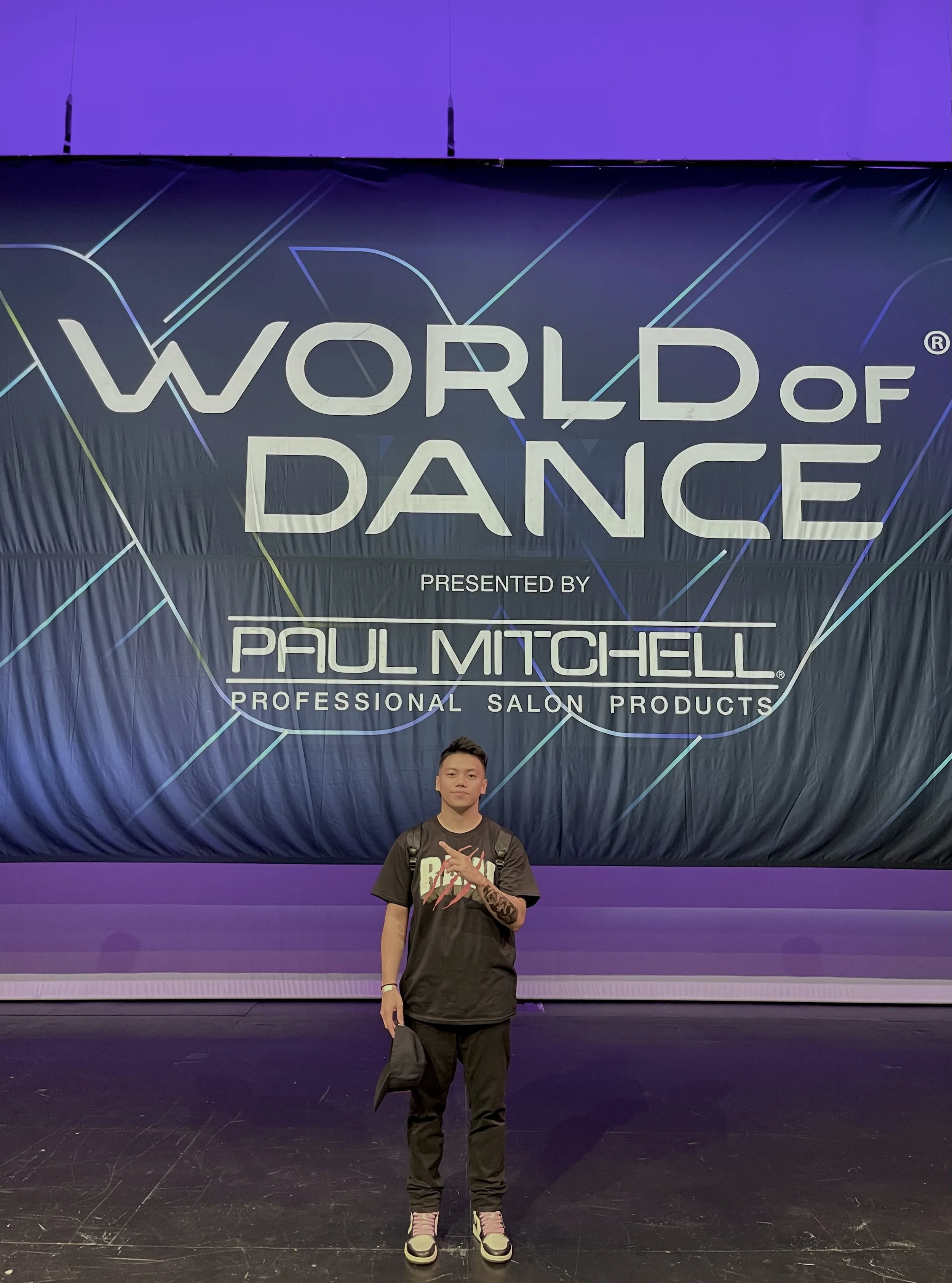 Person standing in front of a large screen at the World of Dance event. The screen displays the event name and sponsors, including Paul Mitchell. The person wears a black t-shirt, black pants, and sneakers, and carries a black backpack.