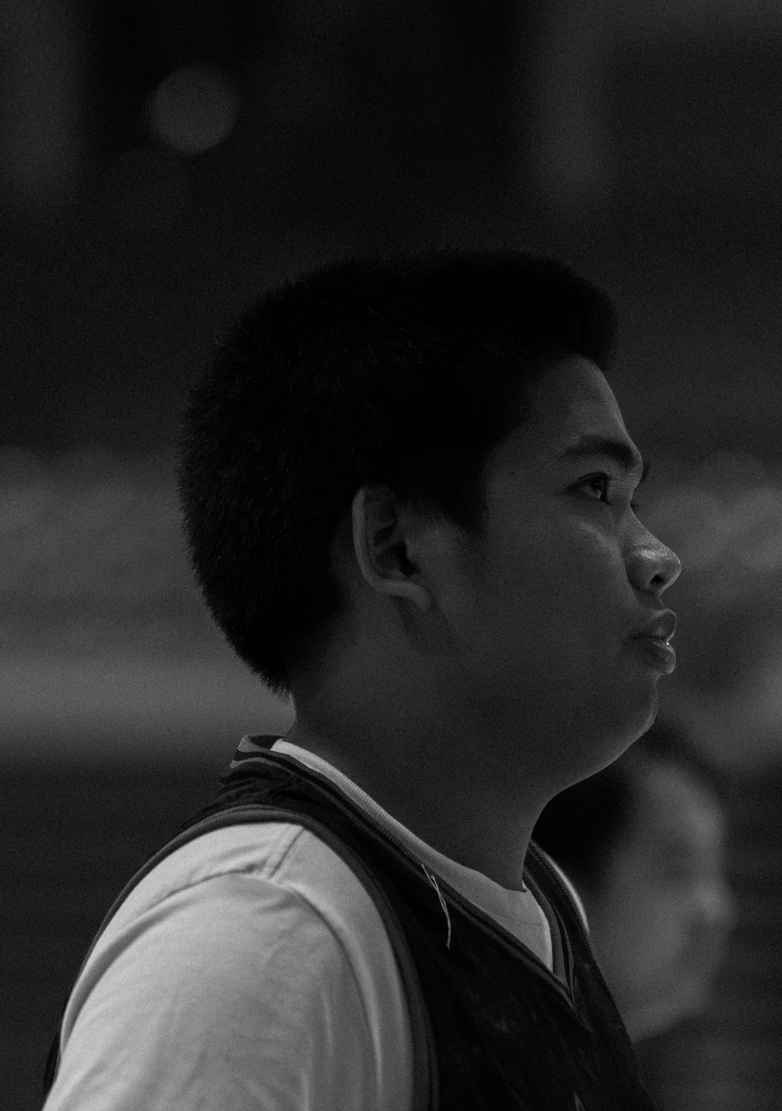 A young man with short hair in profile view, wearing a shirt and vest, standing outdoors at dusk or night, with a dark cloudy sky in the background.
