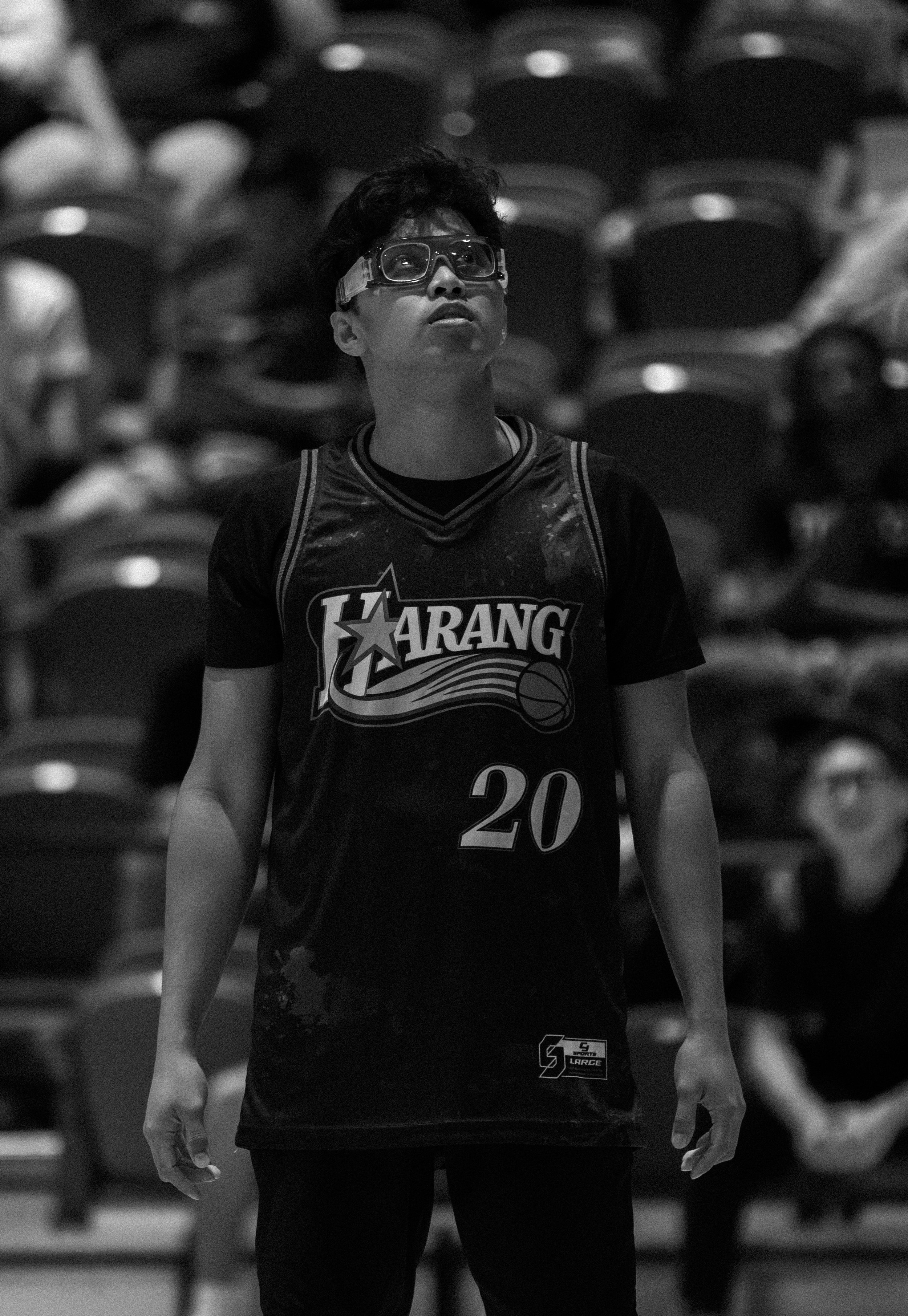 A young man wearing a basketball jersey with the number 20, glasses, and a protective face shield, looking upward in an indoor stadium with empty seats in the background.