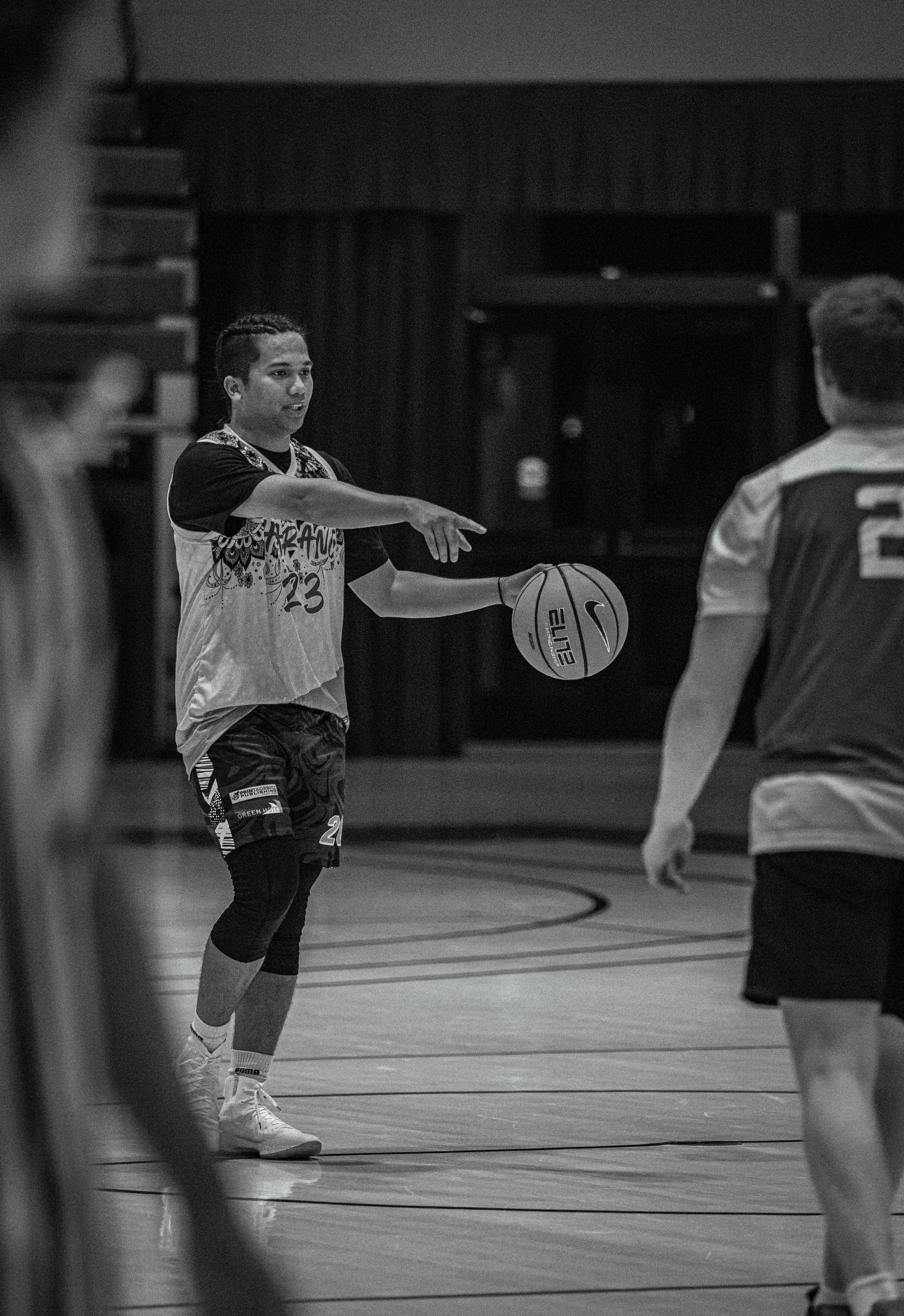 A young male basketball player with braided hair holding a basketball and pointing during a game in an indoor gymnasium.