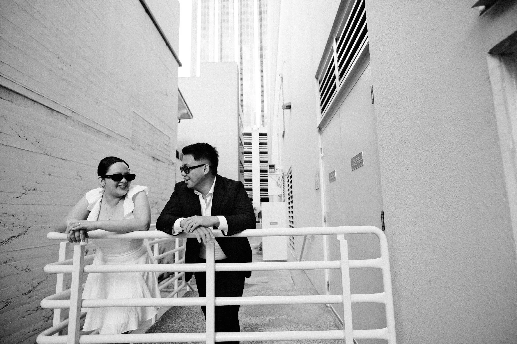 A black and white photo of a woman and a man leaning on a white railing, smiling at each other in an alleyway between modern buildings. The woman is wearing sunglasses and a dress, while the man is in a suit and sunglasses.