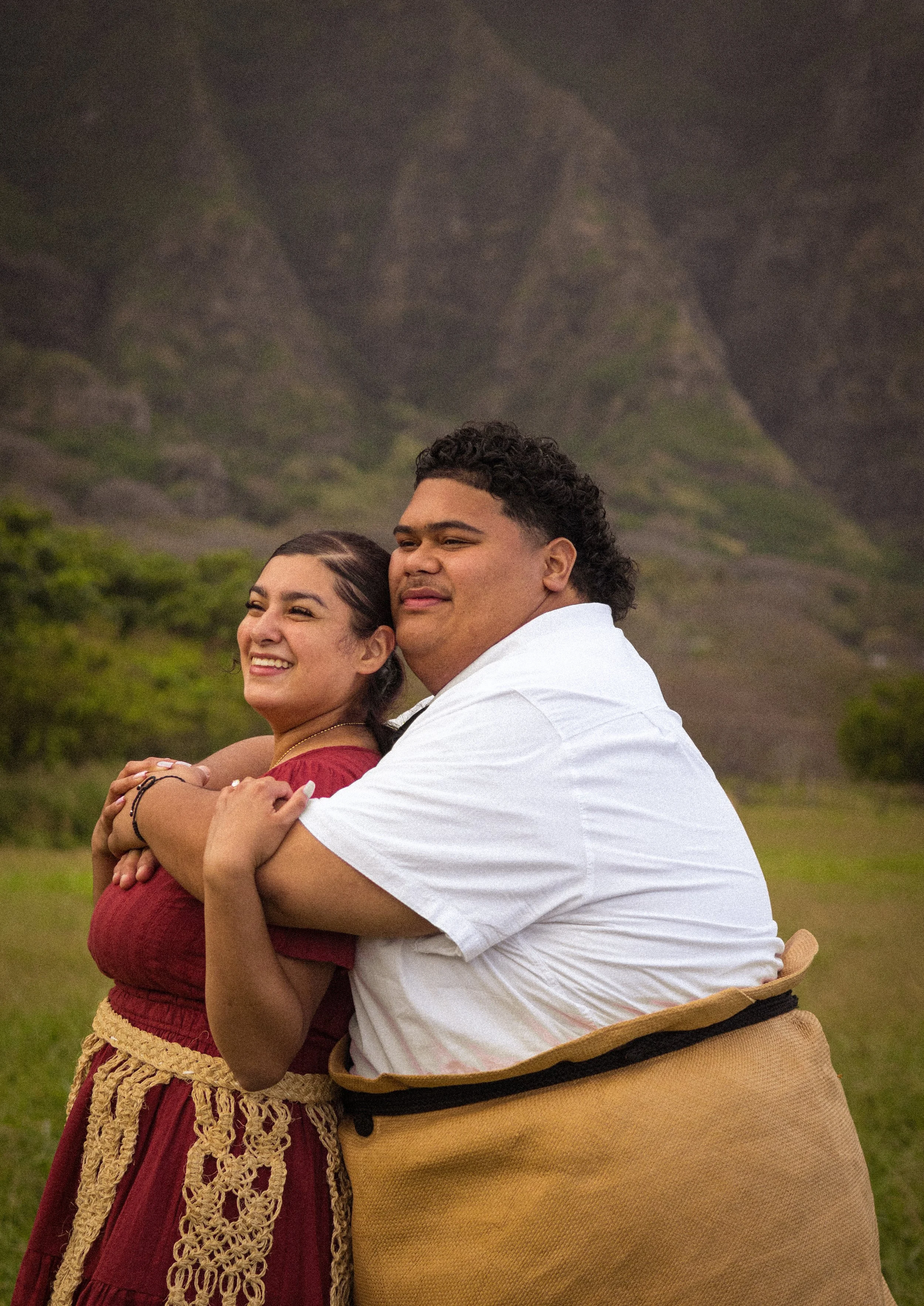 A smiling couple embracing outdoors with mountains in the background.