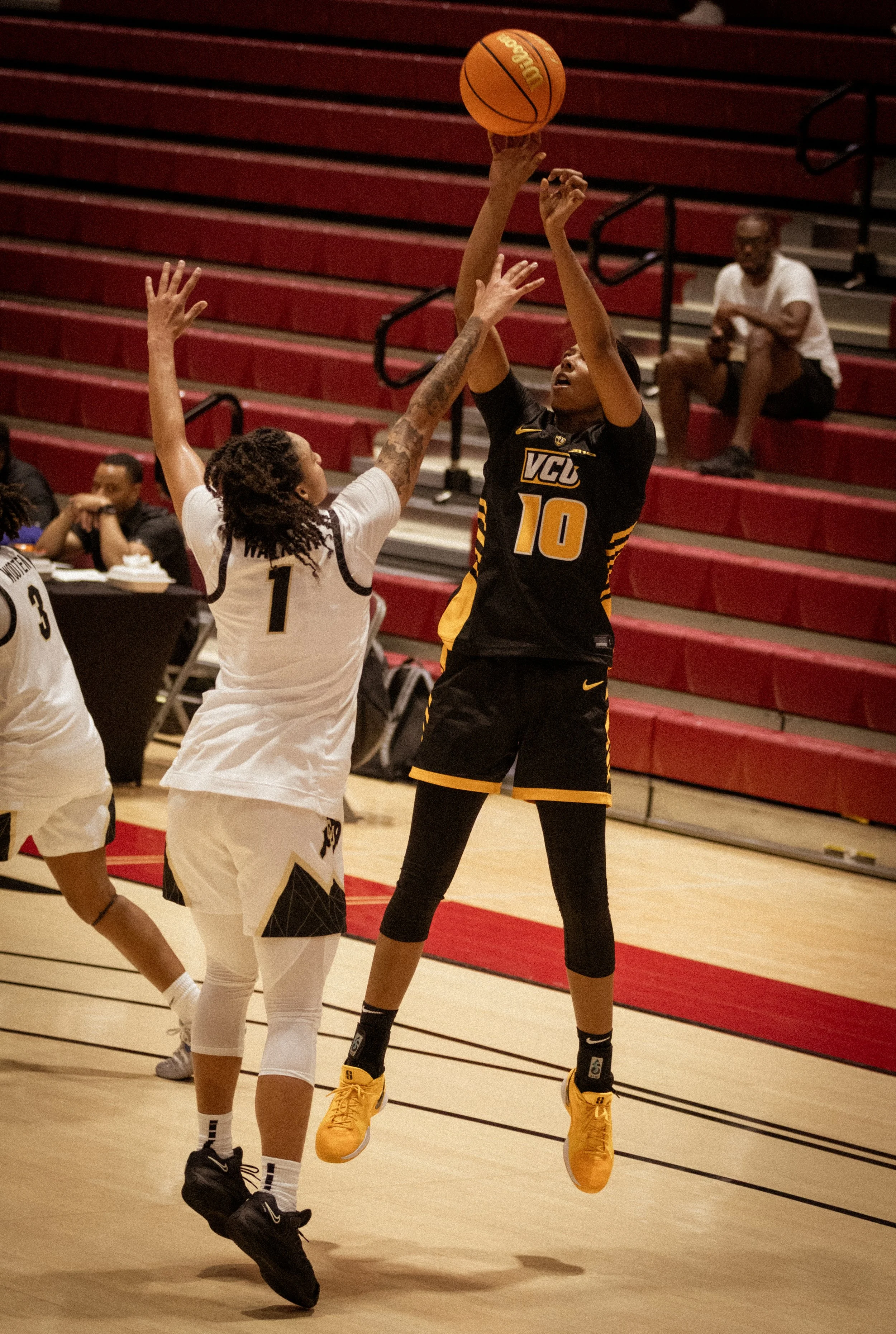 A female volleyball player in a black and yellow uniform jumps to hit the ball, while an opposing player in a white uniform reaches up to block.