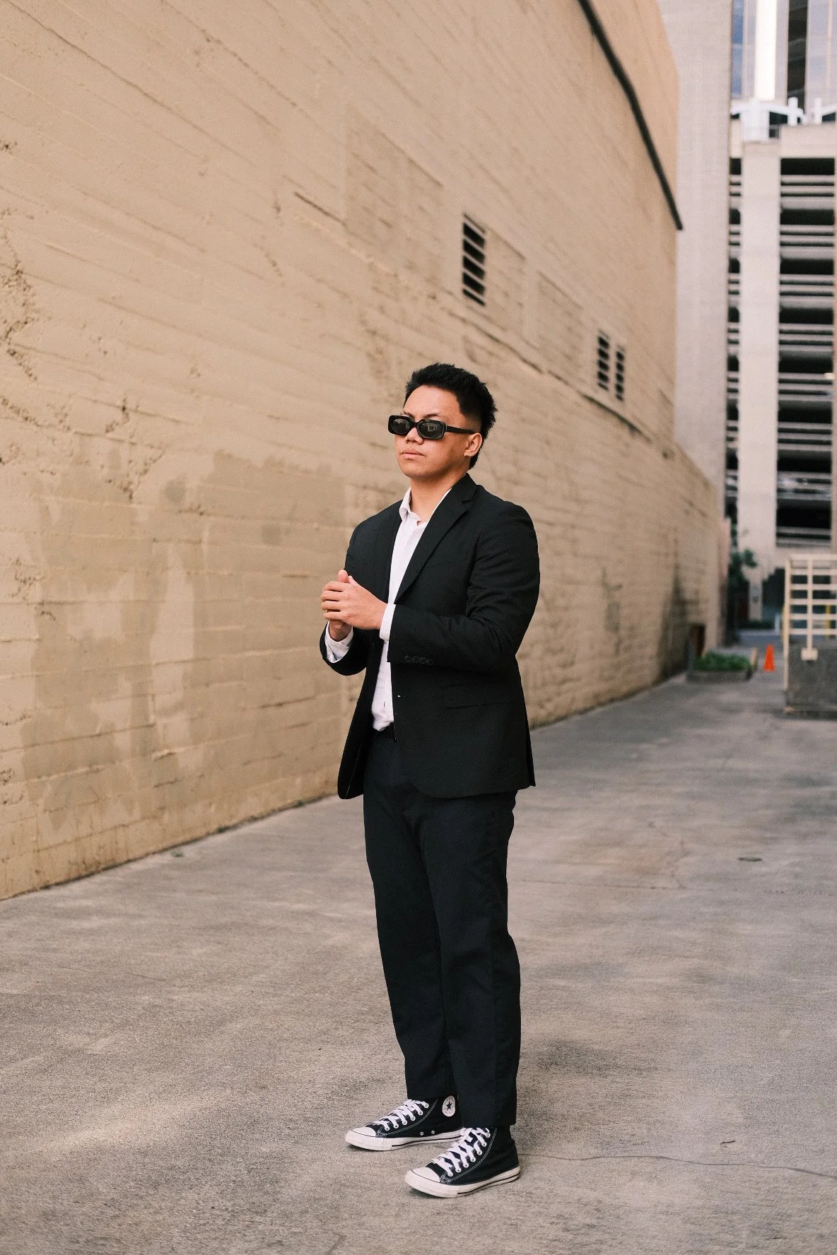 A young man dressed in a black suit and white shirt wearing black sunglasses and black Converse sneakers standing against a beige brick wall in an urban alleyway.
