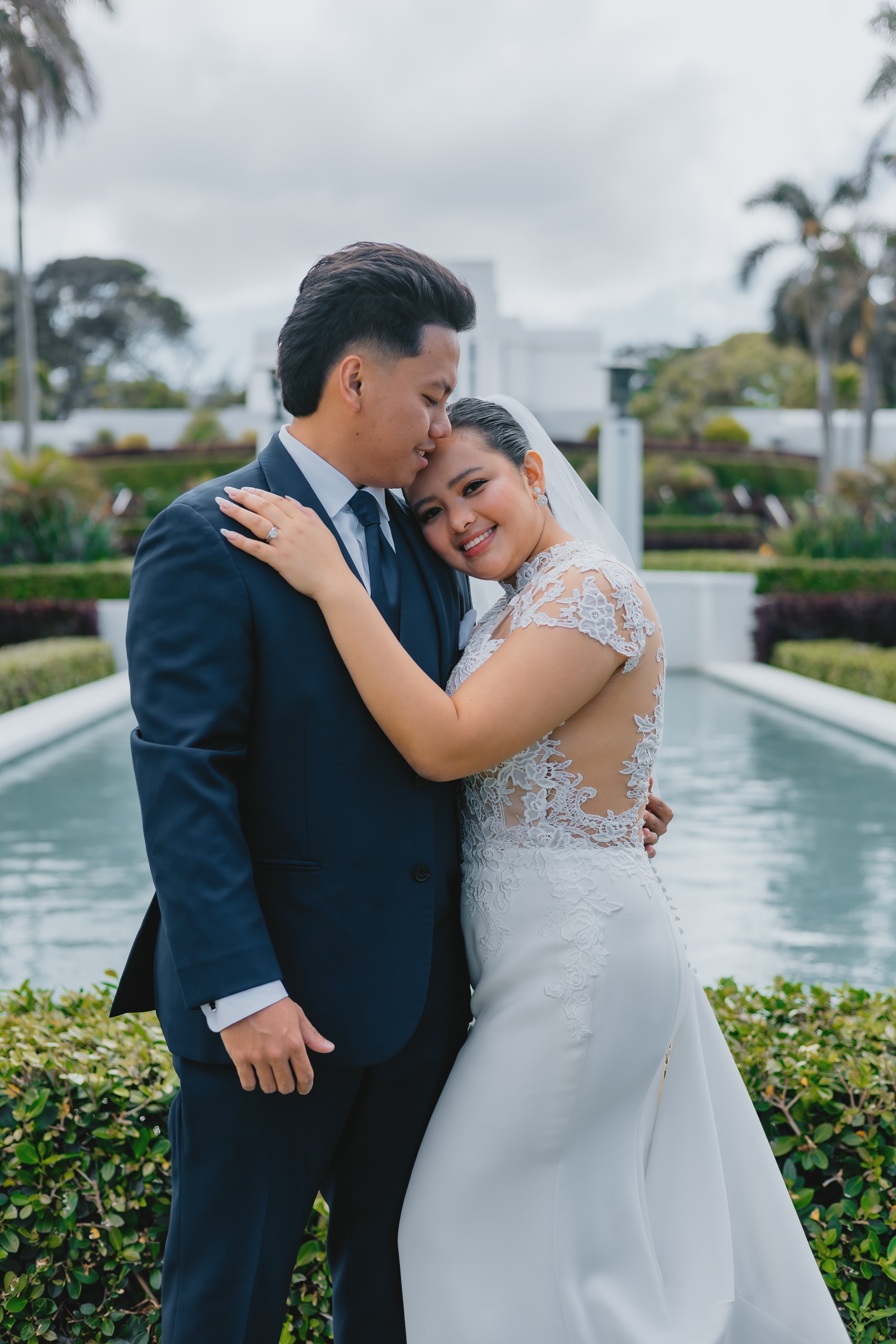 A newlywed couple embracing outdoors by a water feature, with trees and buildings in the background.