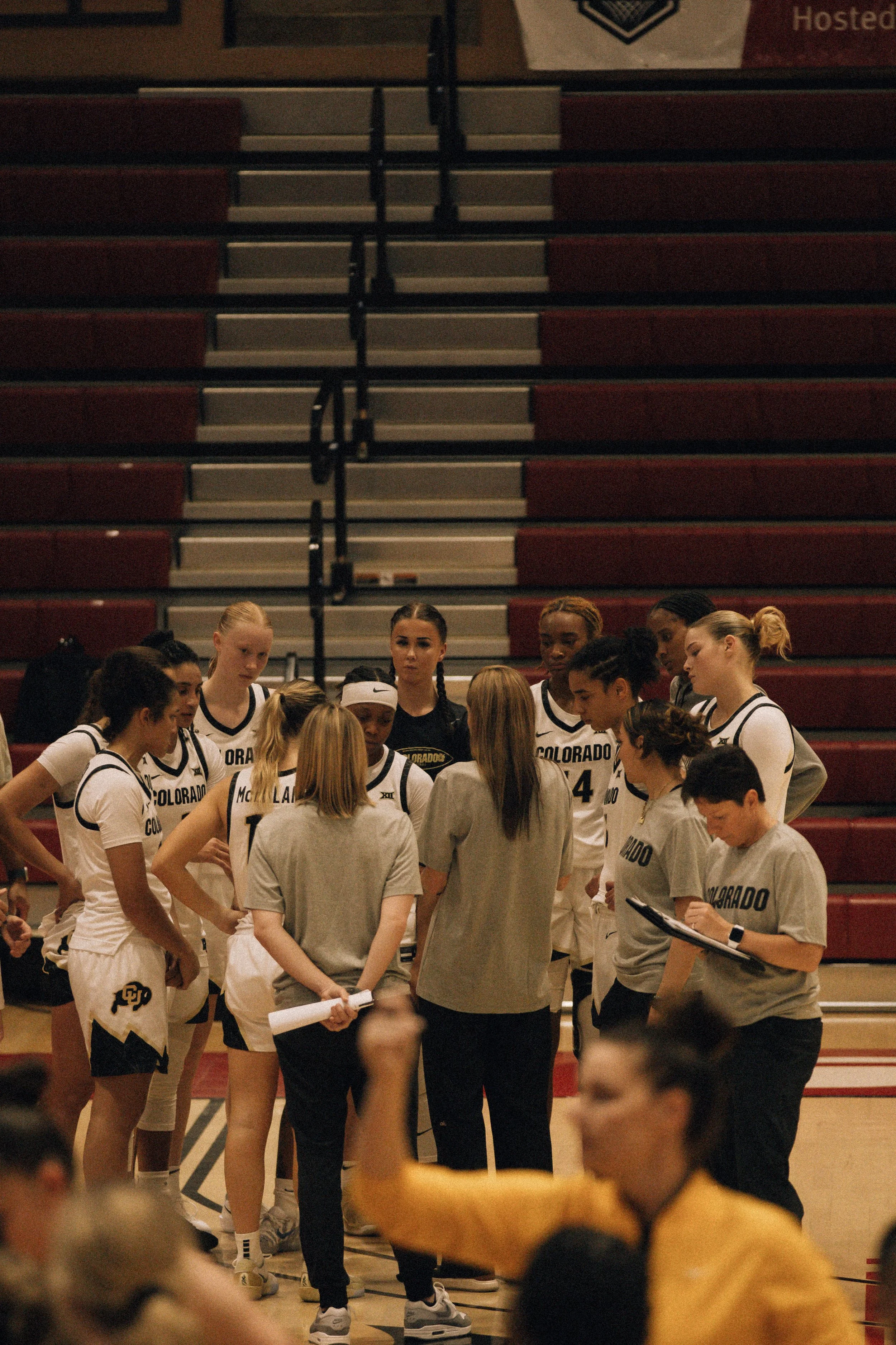A women's basketball team in uniforms listening to their coach during a timeout in a gymnasium, with a blurred person in the foreground demonstrating a fist gesture.