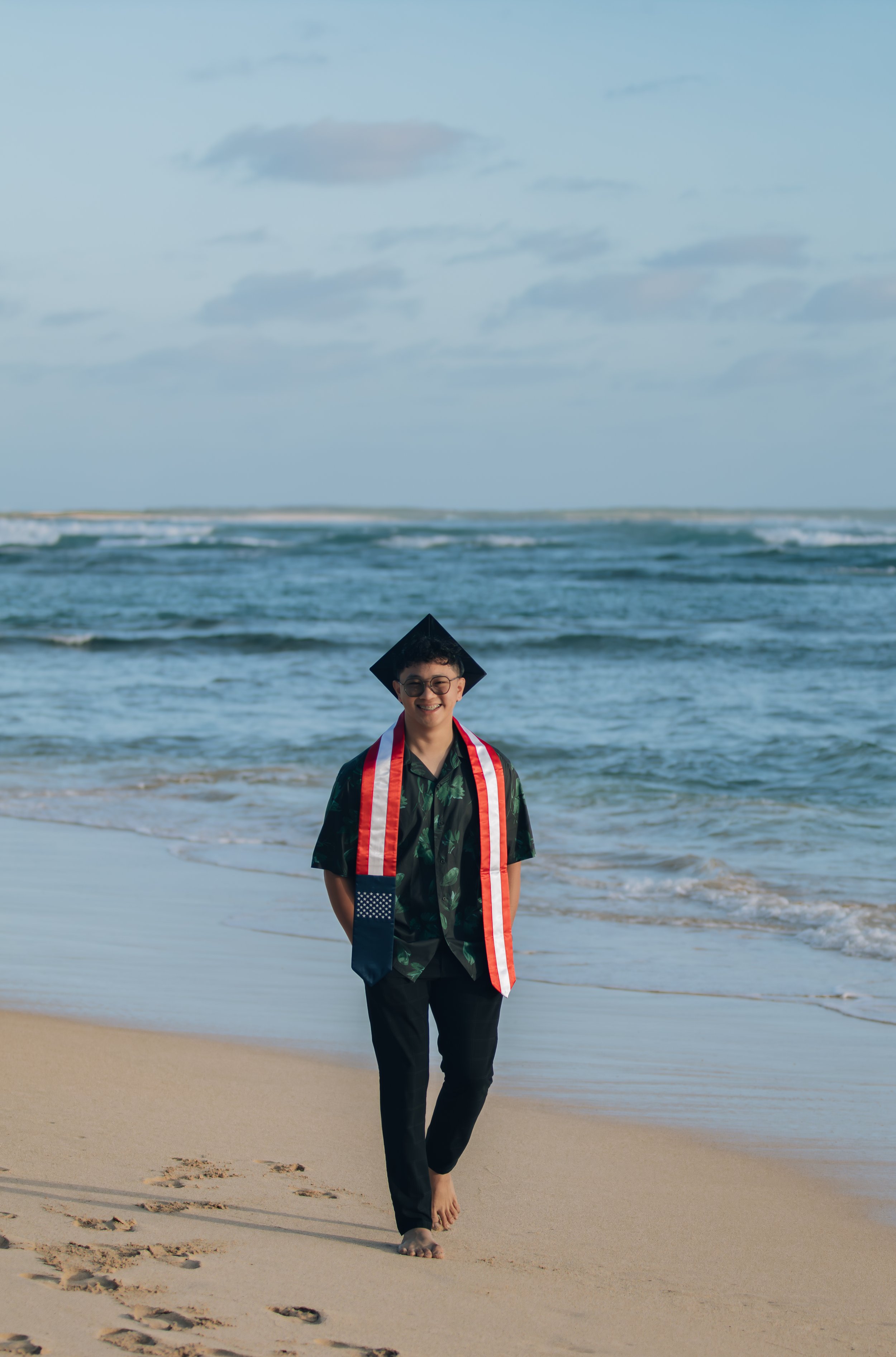 A young man wearing a graduation cap, diploma sash, green tropical shirt, and dark pants walking barefoot on the beach with the ocean in the background.