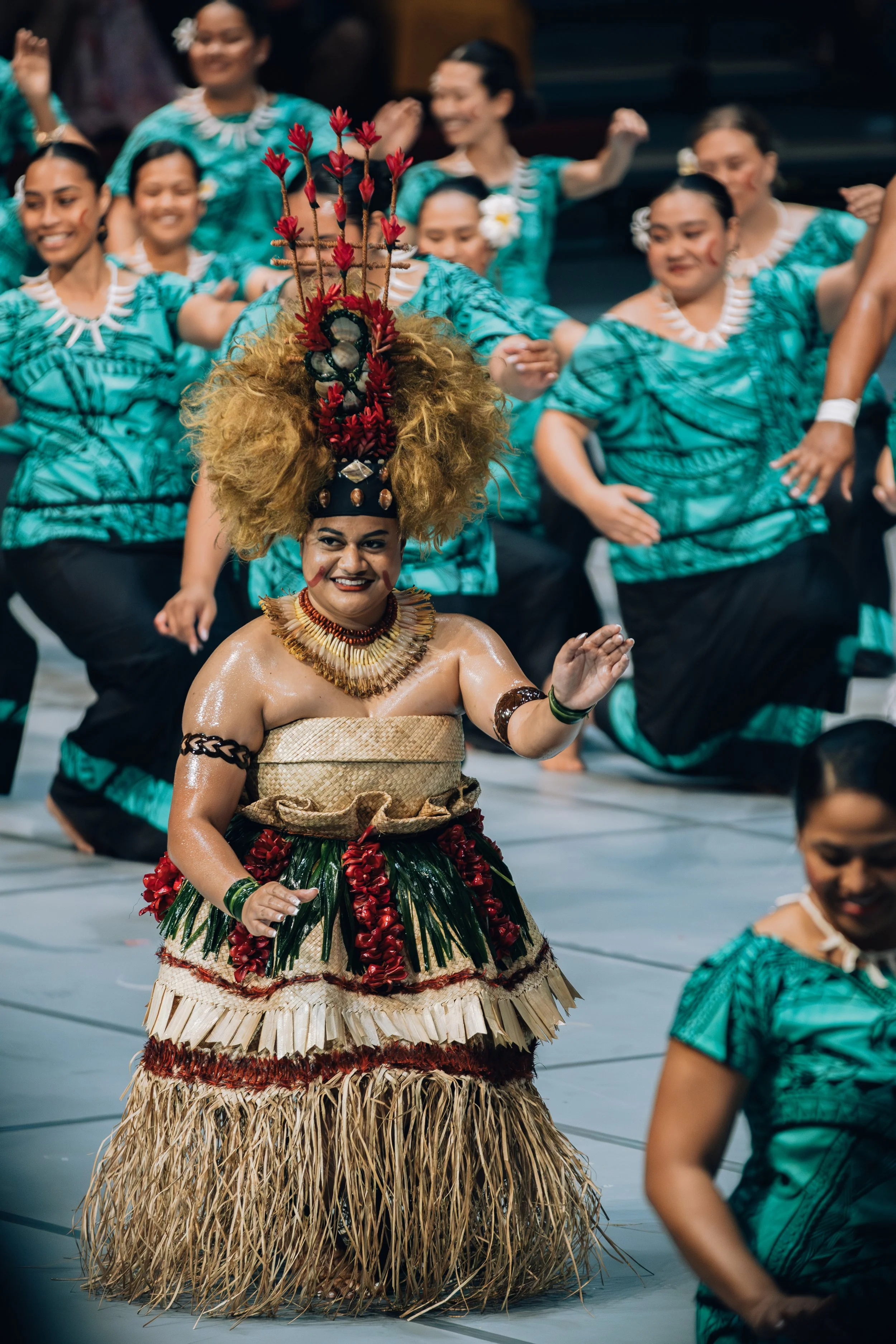 Woman in traditional Polynesian attire, including a grass skirt and elaborate headpiece, performing a dance with a group of women in matching turquoise and black costumes.