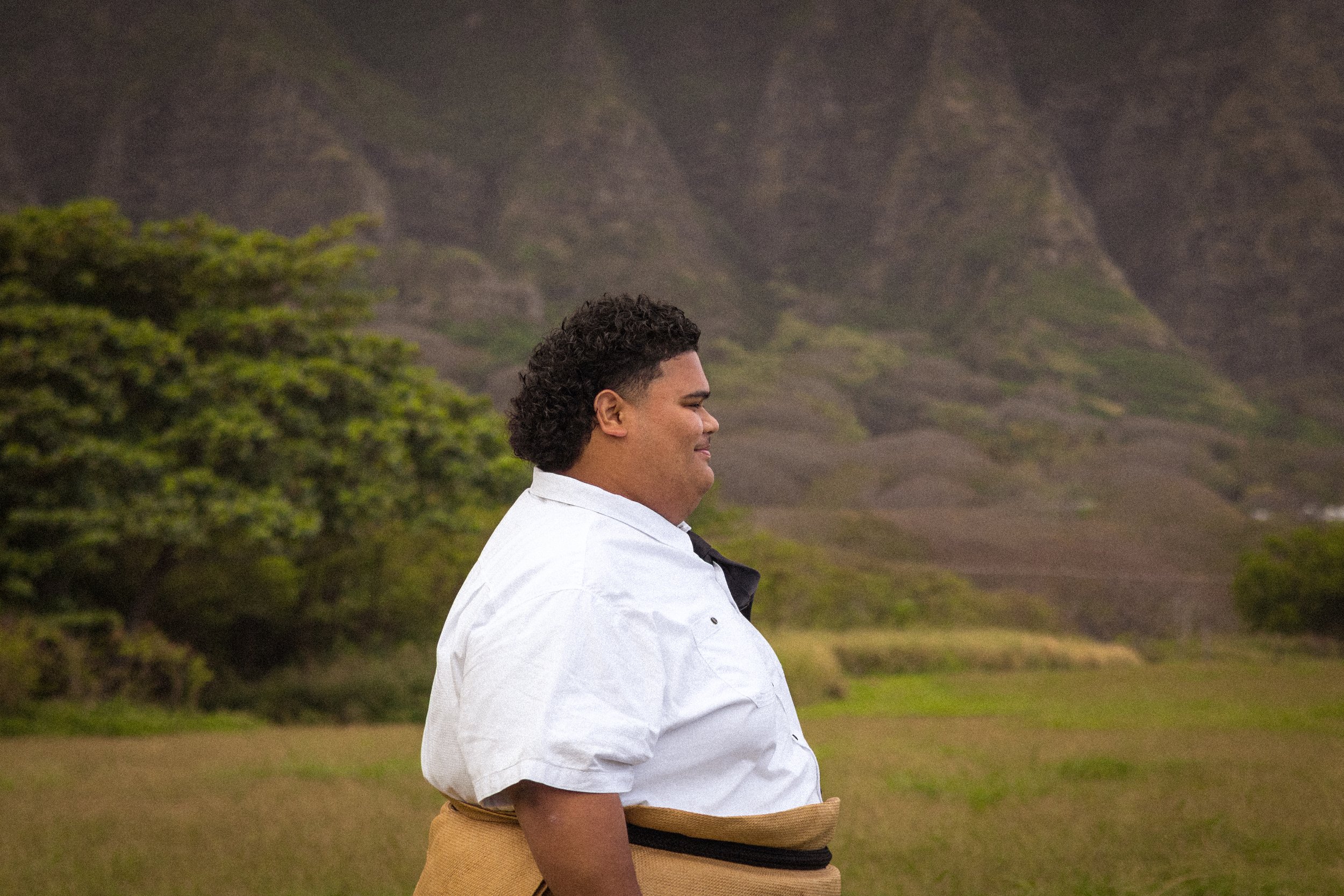 Man in white shirt standing outdoors with mountains and greenery in the background.