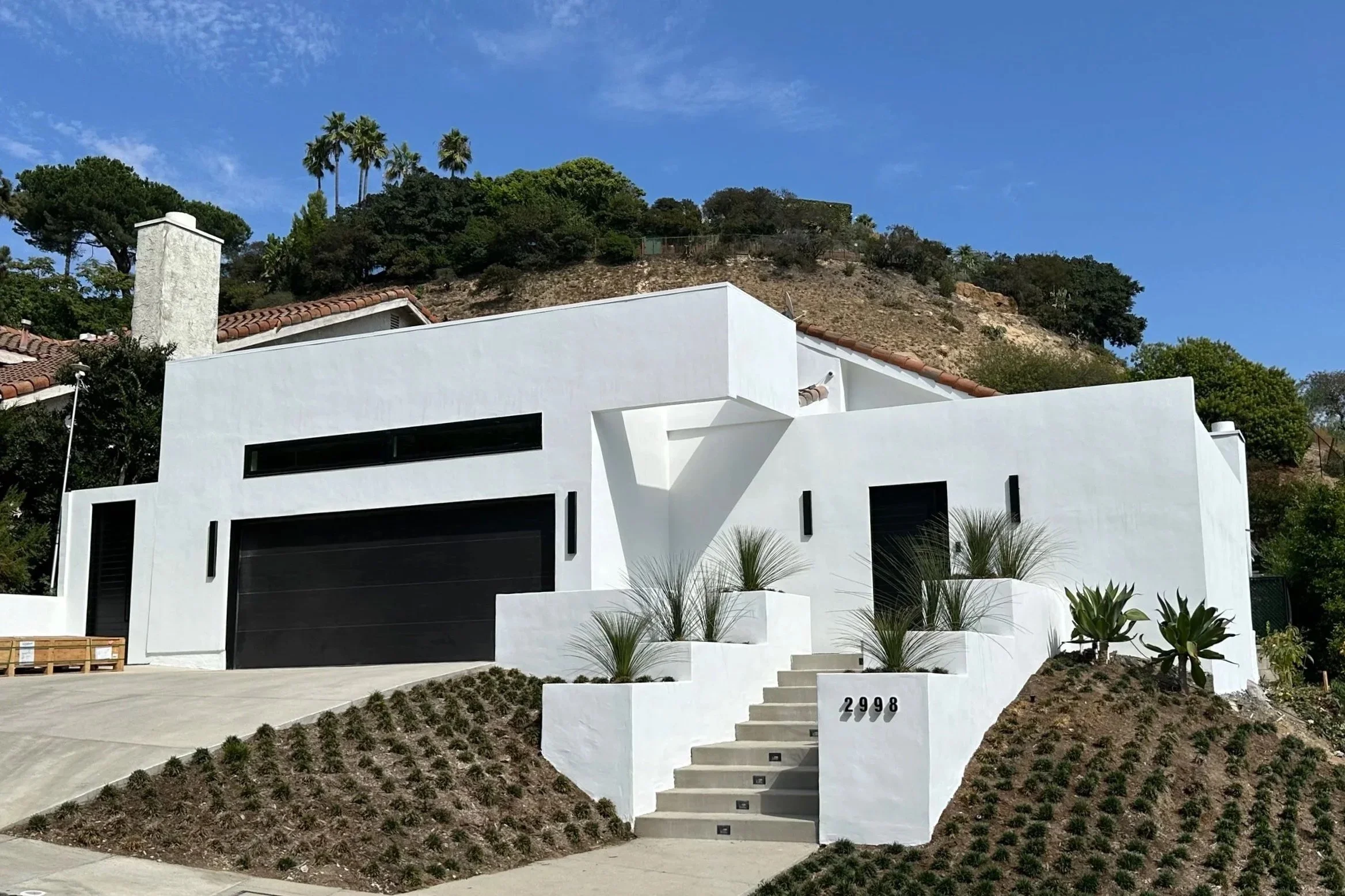 Modern white house with black garage door and minimal landscaping, located on a hillside with a blue sky above.