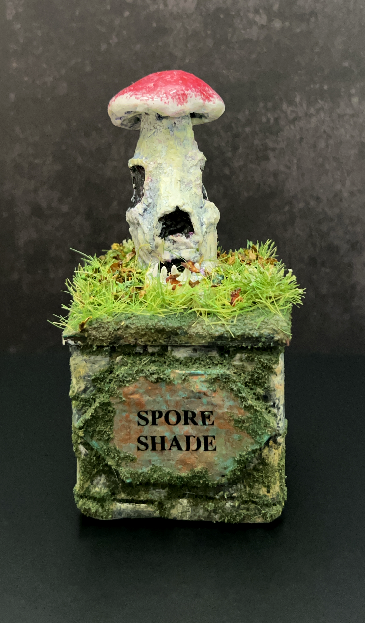 A miniature sculpture of a mushroom with a red cap and a skeletal face on the stem, surrounded by green moss and grass, in front of a rectangular base with a sign that reads 'Spore Shade'