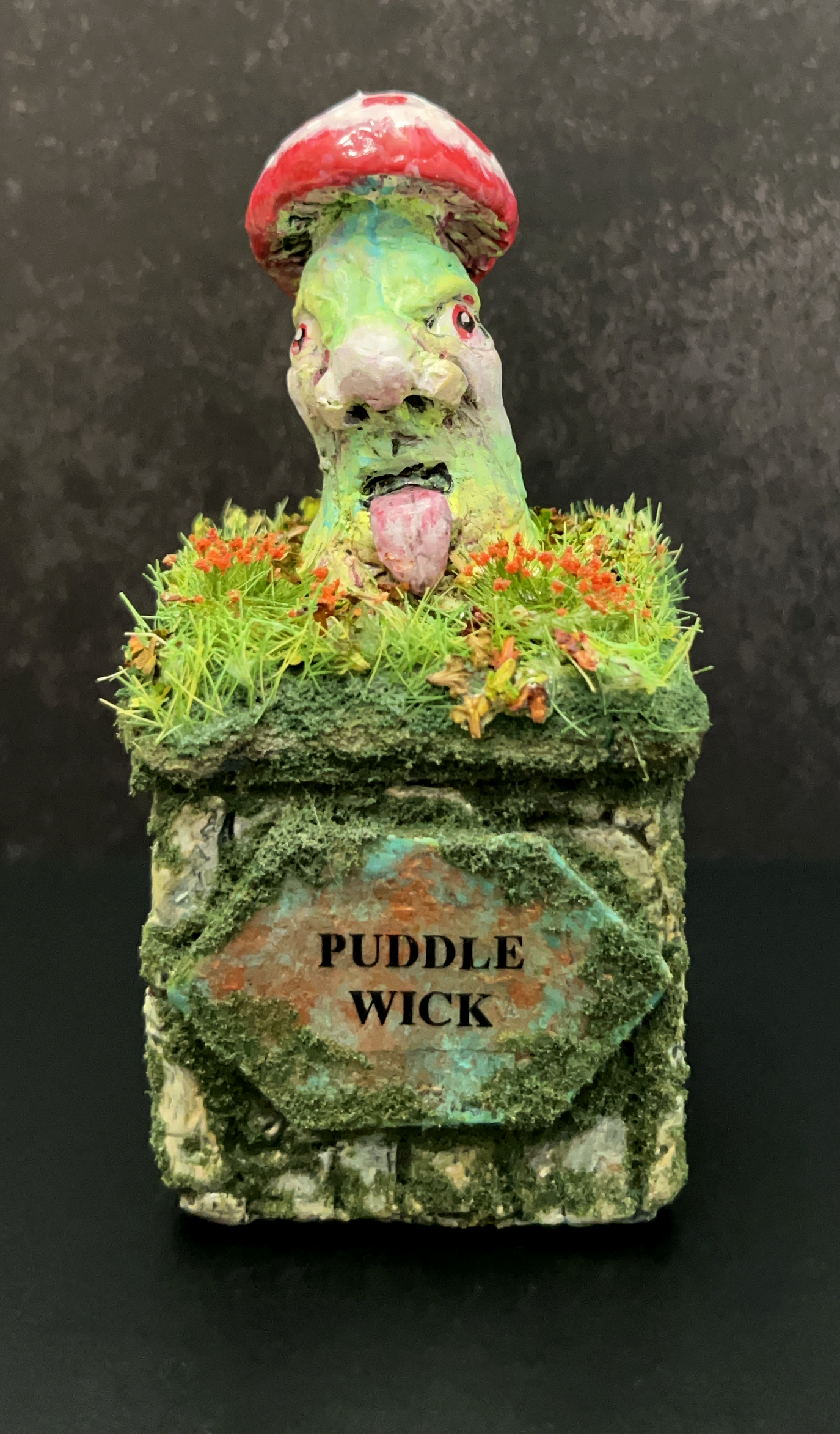 A whimsical garden gnome sculpture with a face, sticking out its tongue, wearing a red mushroom hat, and surrounded by small orange flowers and green moss. The base has a sign that reads 'PUDDLE WICK'.