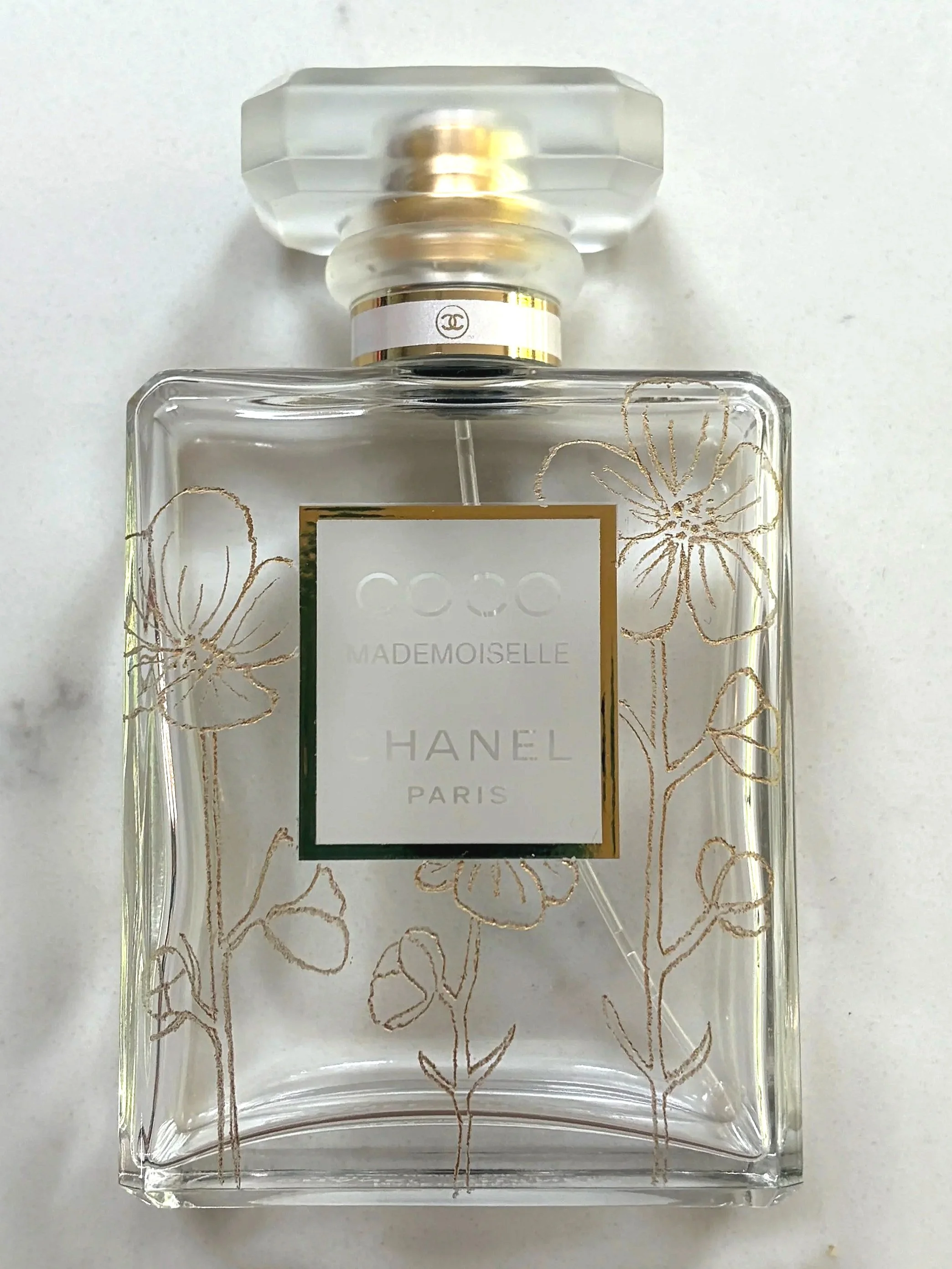 A clear glass Chanel perfume bottle with hand engraved gold floral designs on the front, labeled as Coco Mademoiselle Paris, topped with a transparent hexagonal cap.