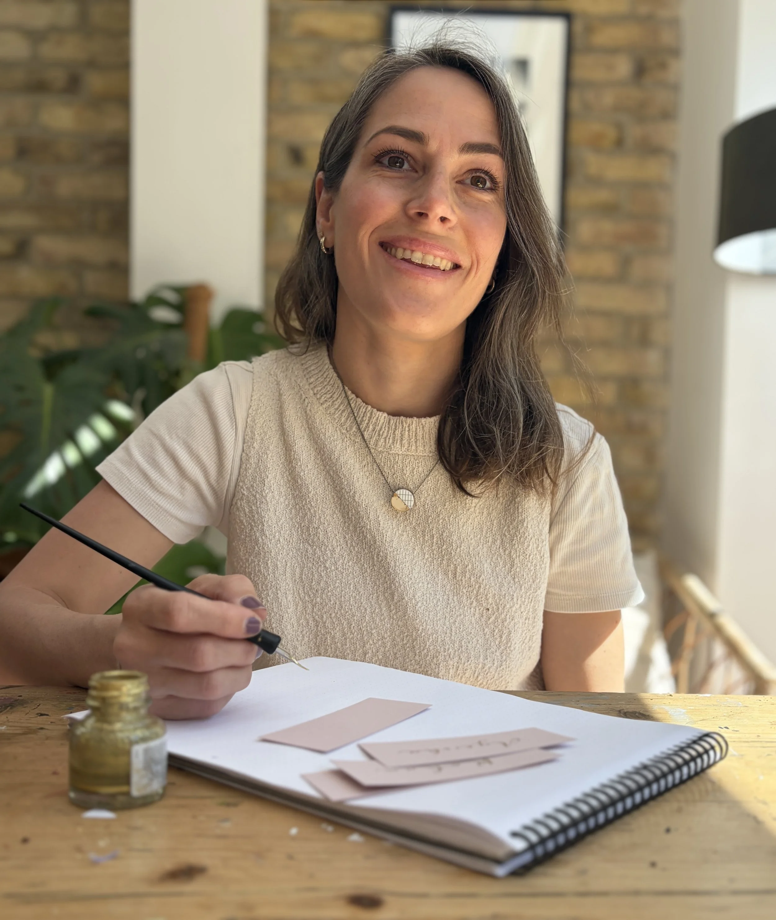 A woman with brown hair sitting at a wooden table, smiling, with calligraphy supplies and paper in front of her