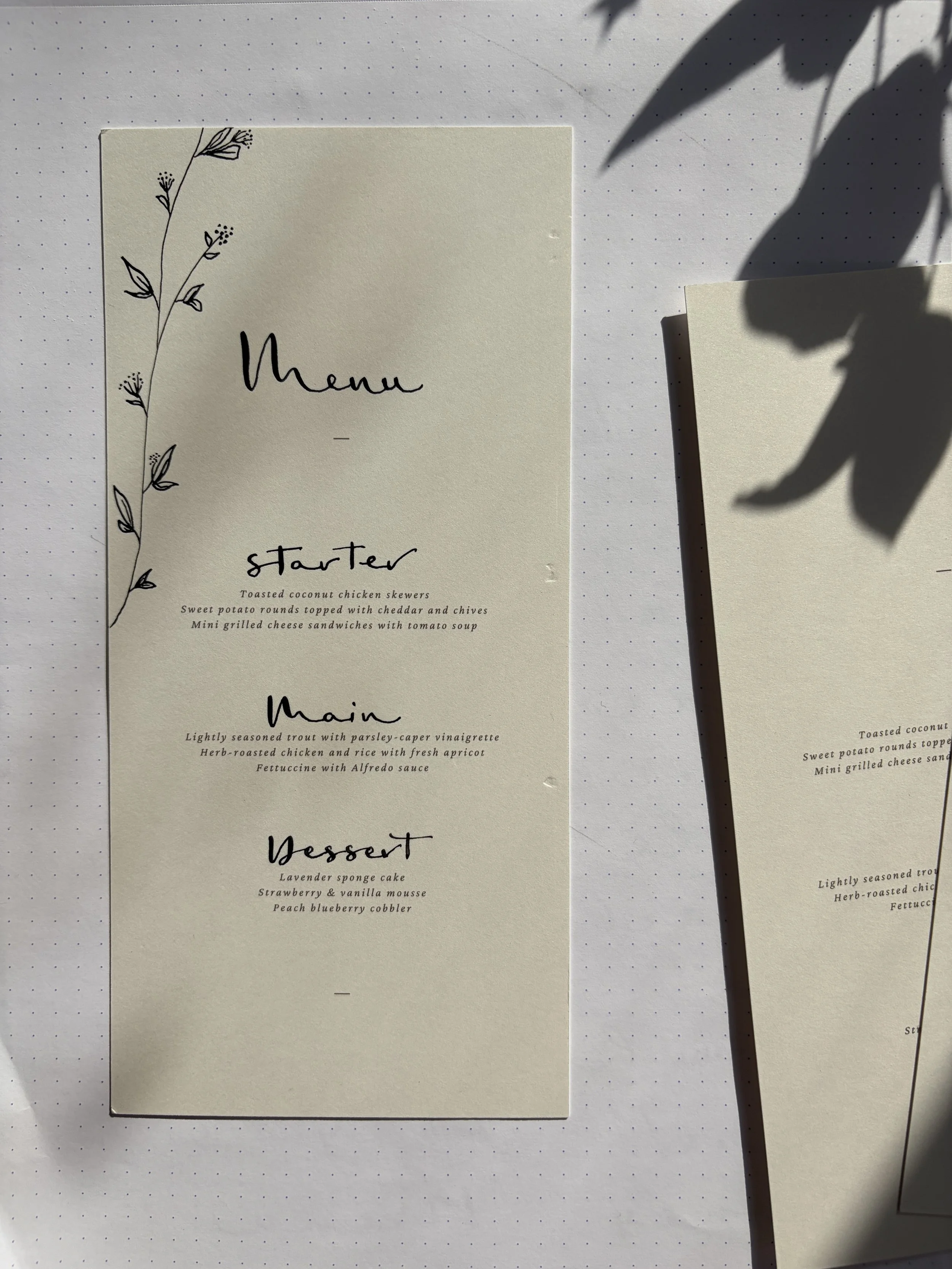 A menu card with a hand-drawn floral illustration on the left side, listing three sections: "starter," "main," and "dessert," with descriptions of various dishes below each section heading. Part of another similar menu is visible to the right, and th