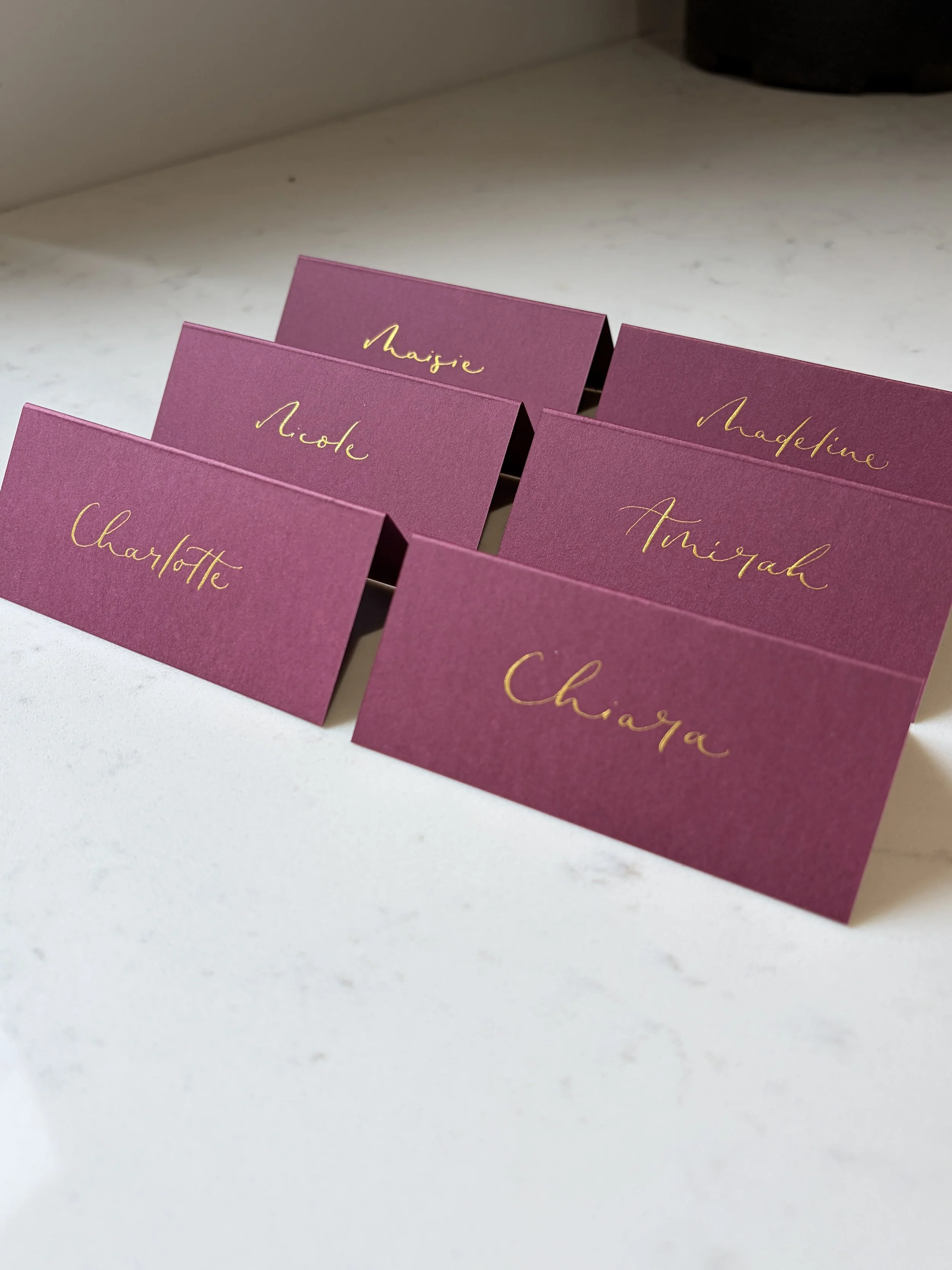 	
Burgundy coloured place cards with gold calligrpahy, displaying names such as Maisie, Madeleine, Charlotte, and Amirah, arranged on a white surface.