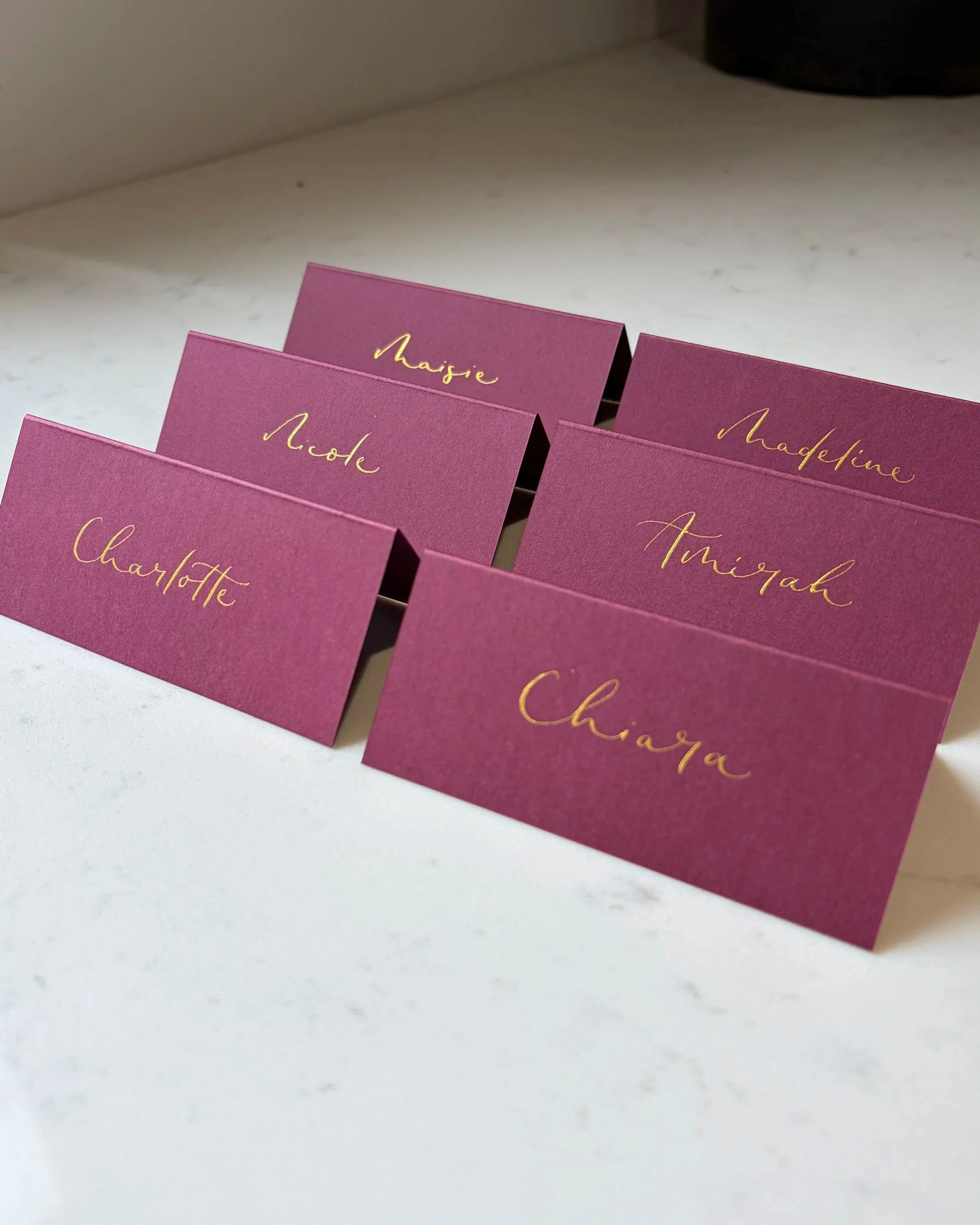 Burgundy coloured place cards with gold calligrpahy, displaying names such as Maisie, Madeleine, Charlotte, and Amirah, arranged on a white surface.