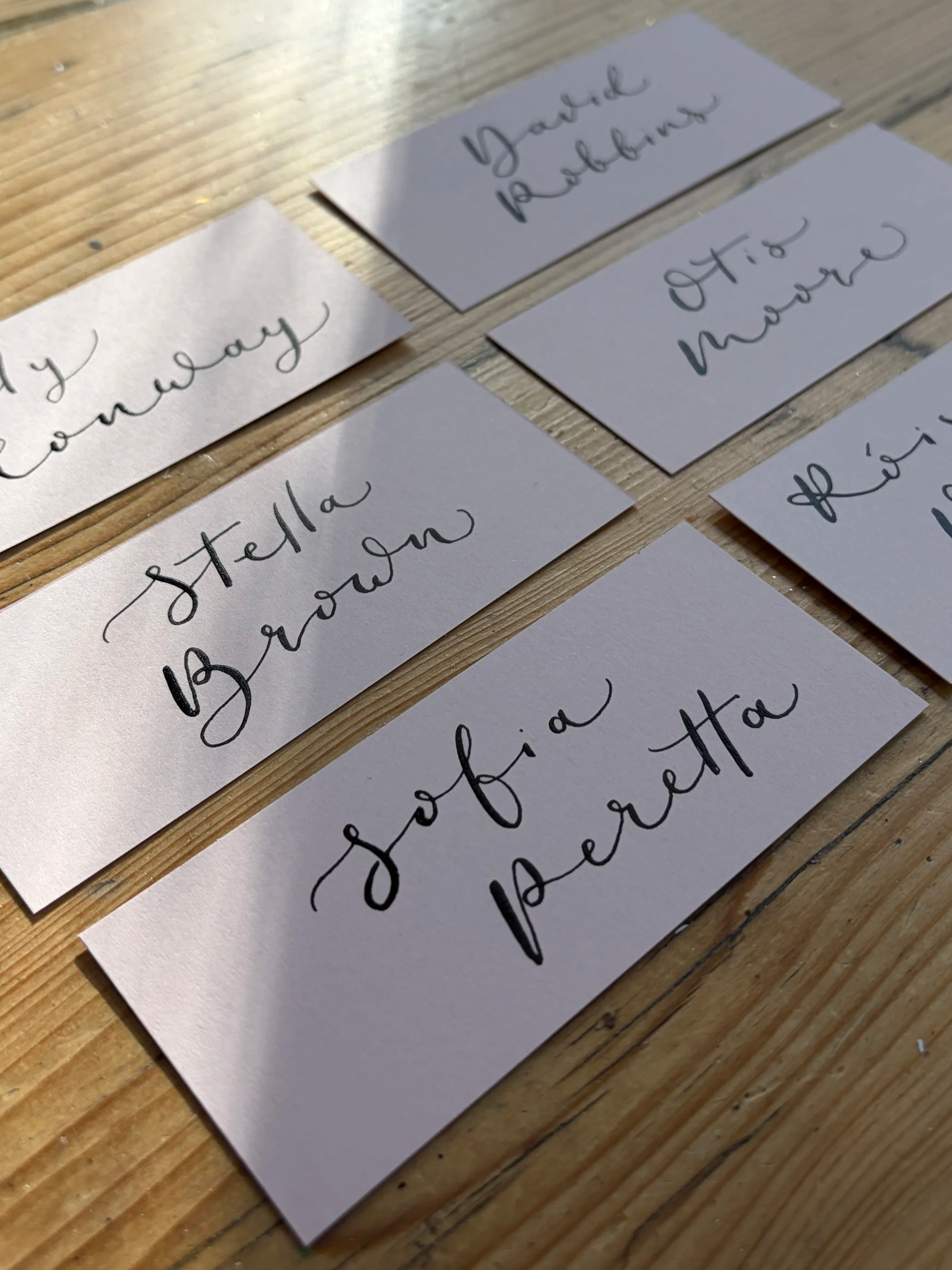 Several pink place cards with names written in black ink calligraphy, laid out on a wooden surface.