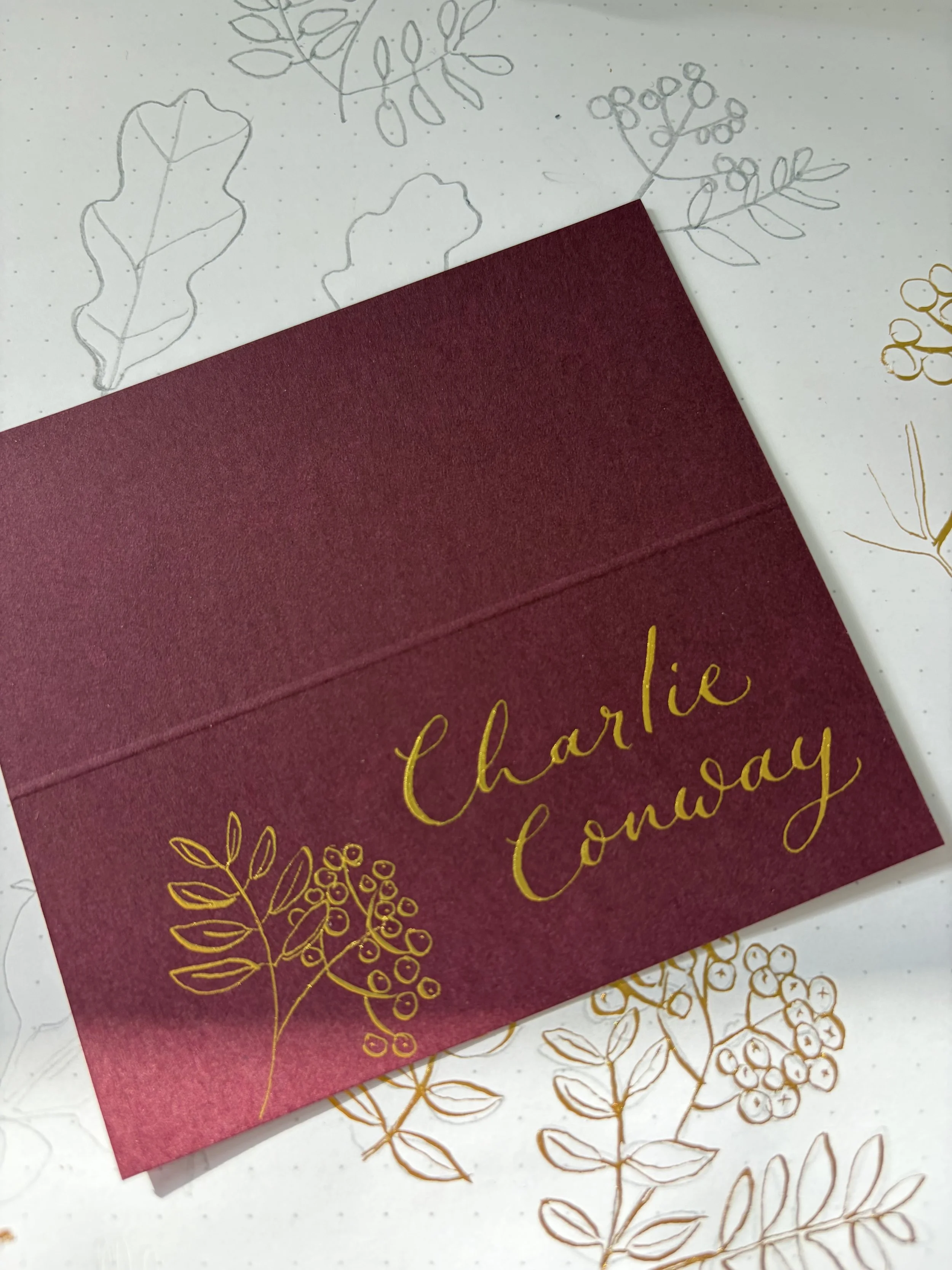 A burgundy coloured placecard with gold calligraphy and gold botanical illustrations on the front. The card is placed on a white background decorated with faint gold botanical drawings behind.