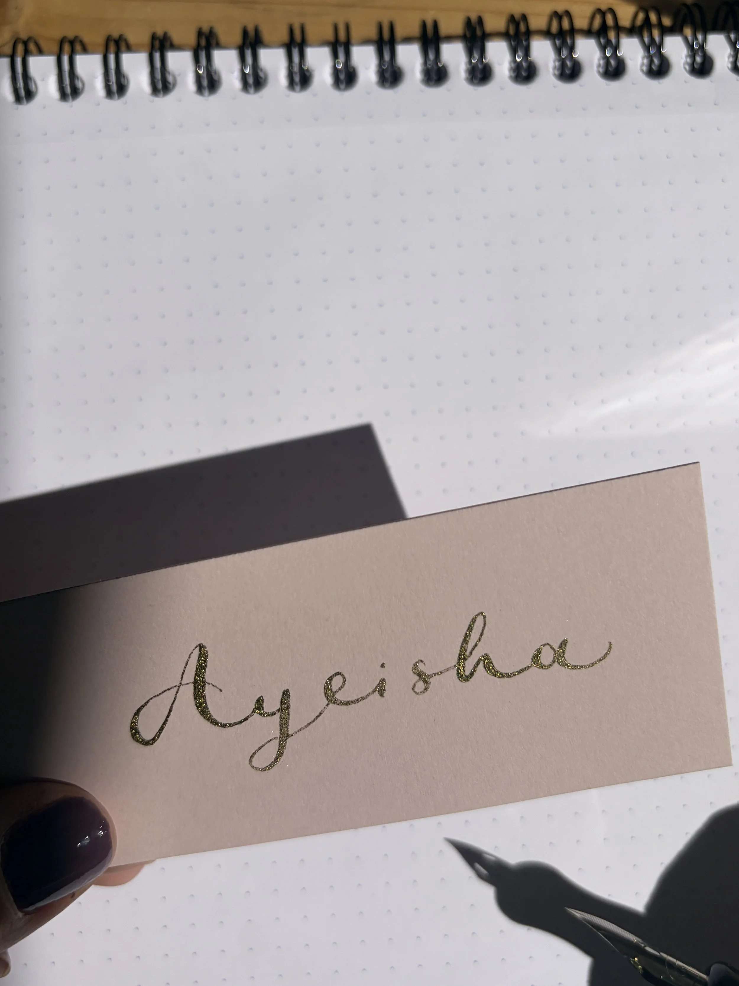 Close-up of a pink placecard with the name Ayeisha written in gold calligraphy. Hand holding a black calligraphy with a metallic nib is visible in the bottom right corner of the photo.