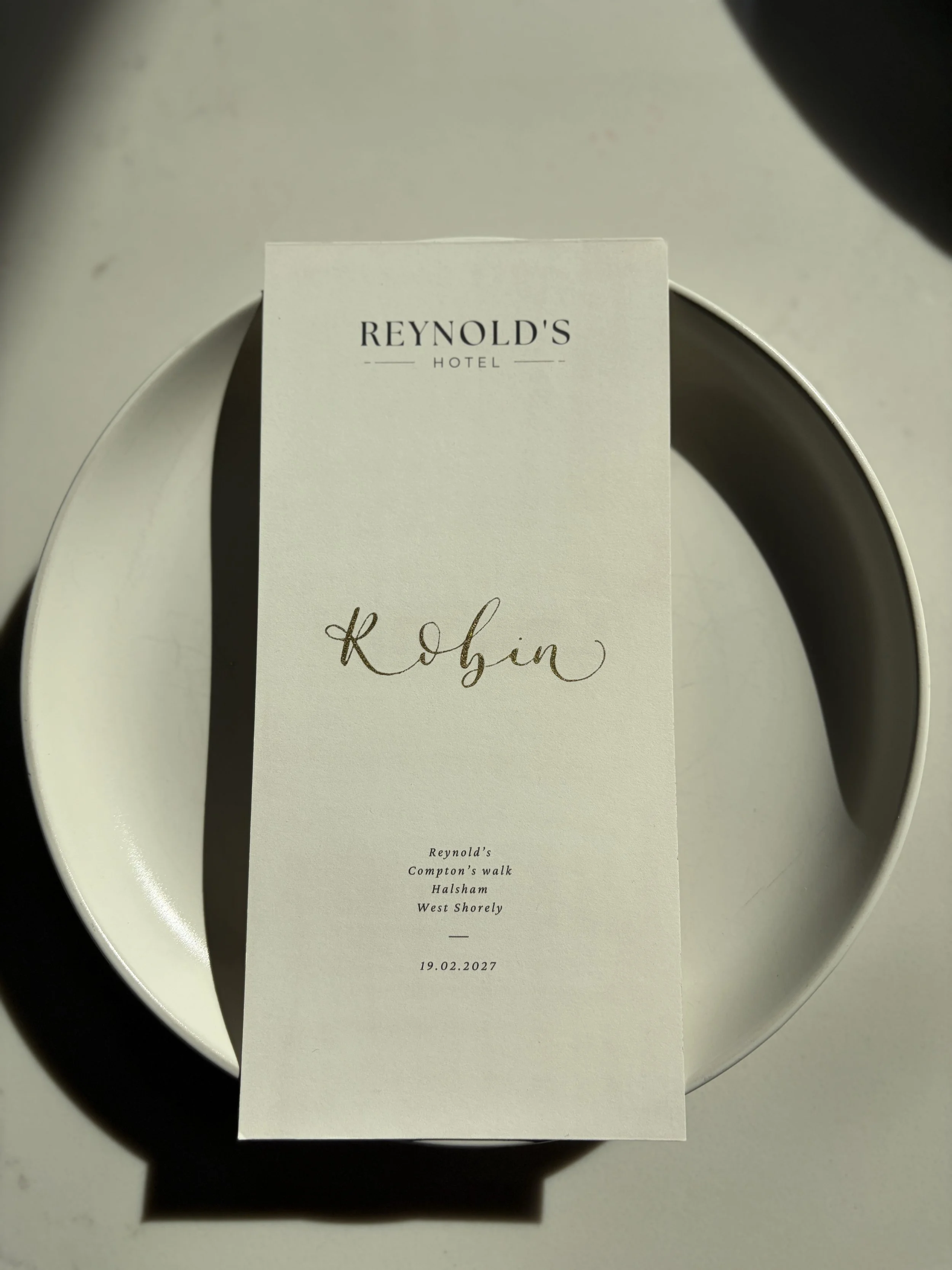A white menu card with the text 'REYNOLD'S HOTEL' at the top, personalised with the name 'Robin' in gold calligraphy. Below that is the location and date, specifically 'Reynold's Compton's walk Halsham West Shorely' and the date '19.02.2027'.
