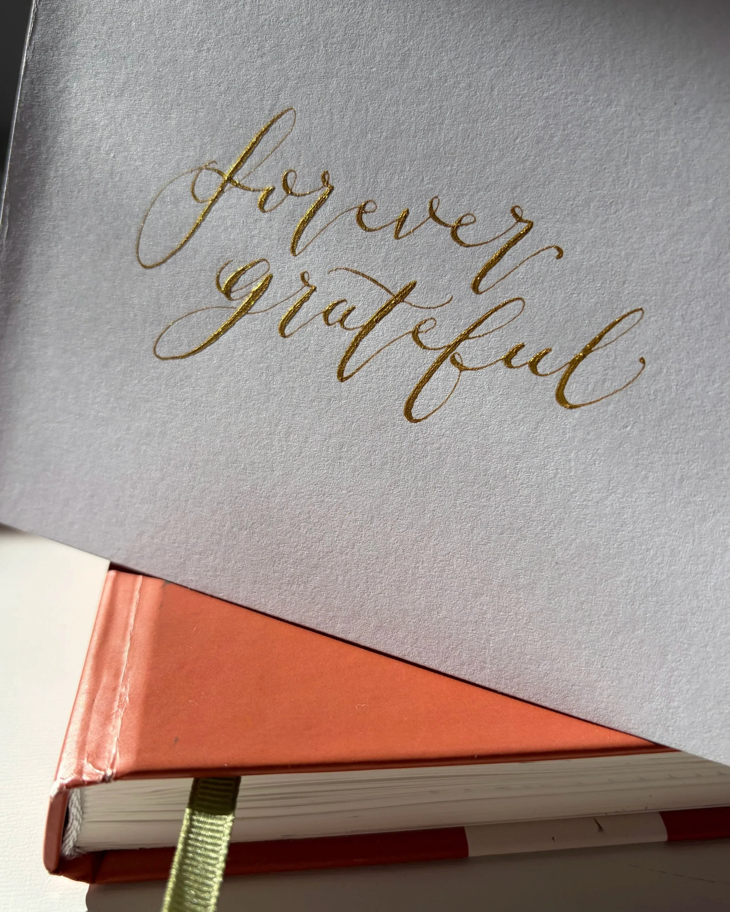 Close-up photo of a  greetings card with gold calligraphy wording "forever grateful". The greetings card is sitting on a closed notebook.