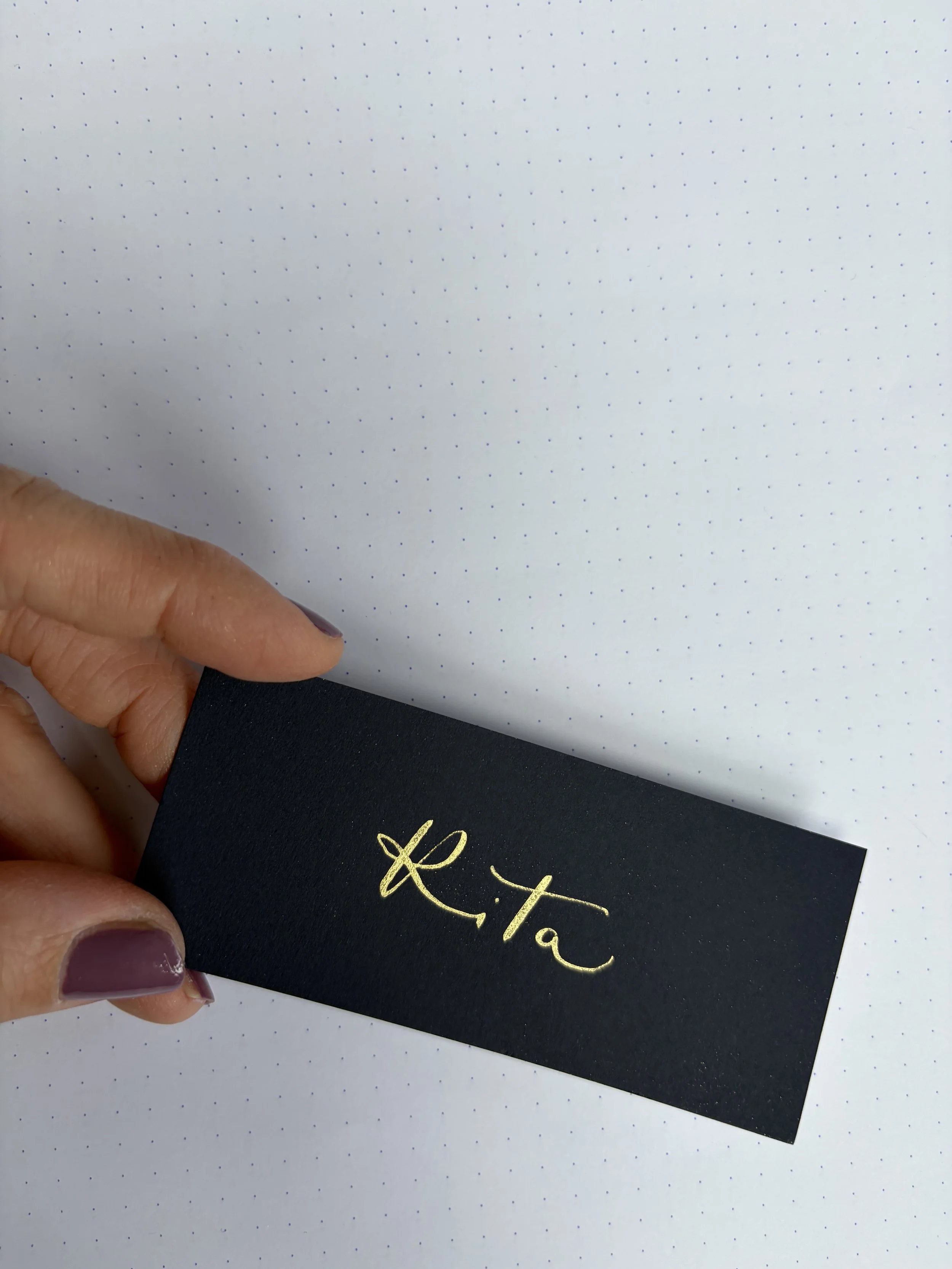 Close-up of a hand holding a dark blue place card with the name 'Rita' written in gold dip-pen calligraphy.
