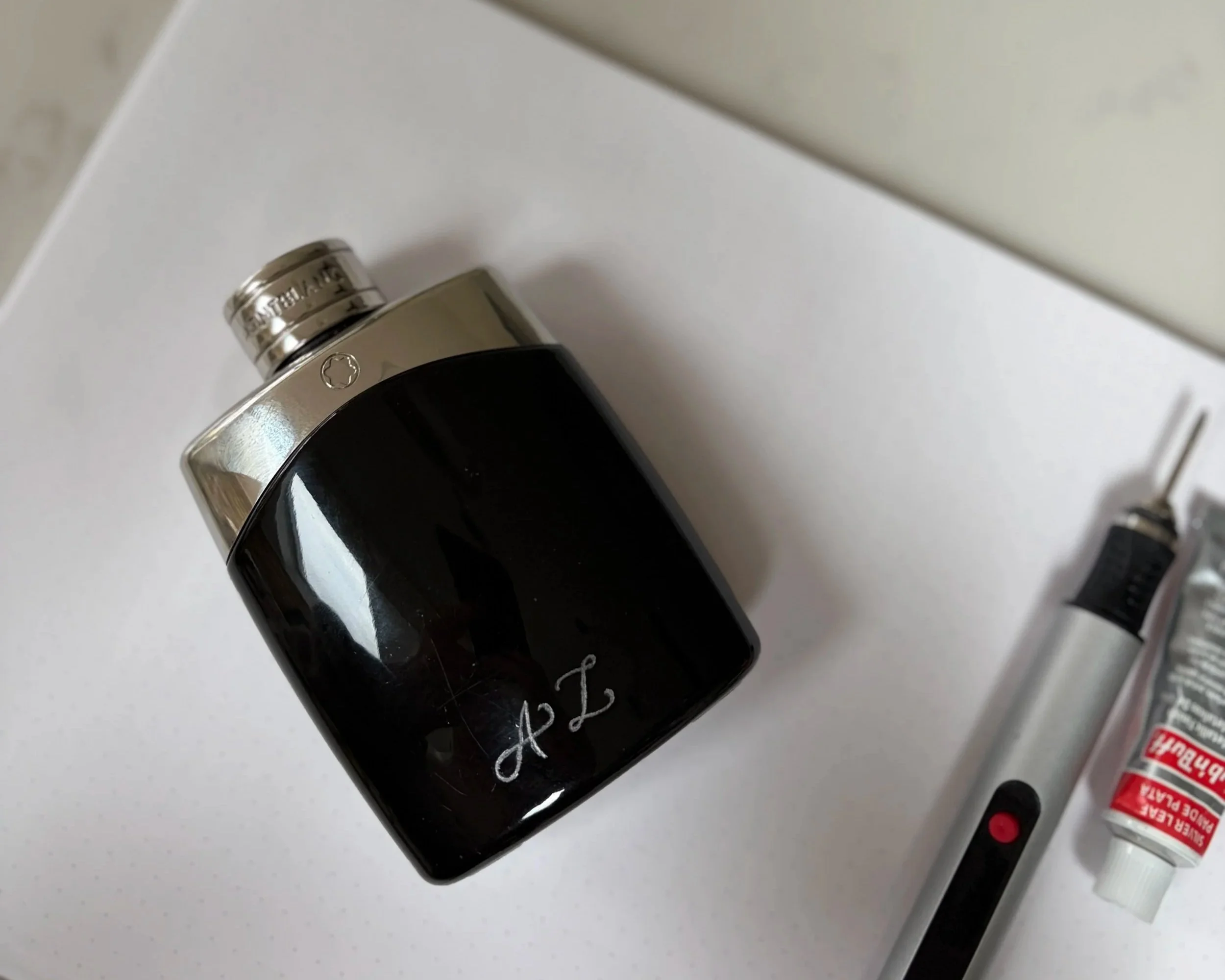 Close-up of a black aftershave bottle with a silver cap resting on a white surface, with handwritten initials engraved on the bottle and embellished in silver. There is an engarving pen and silver paint nearby.