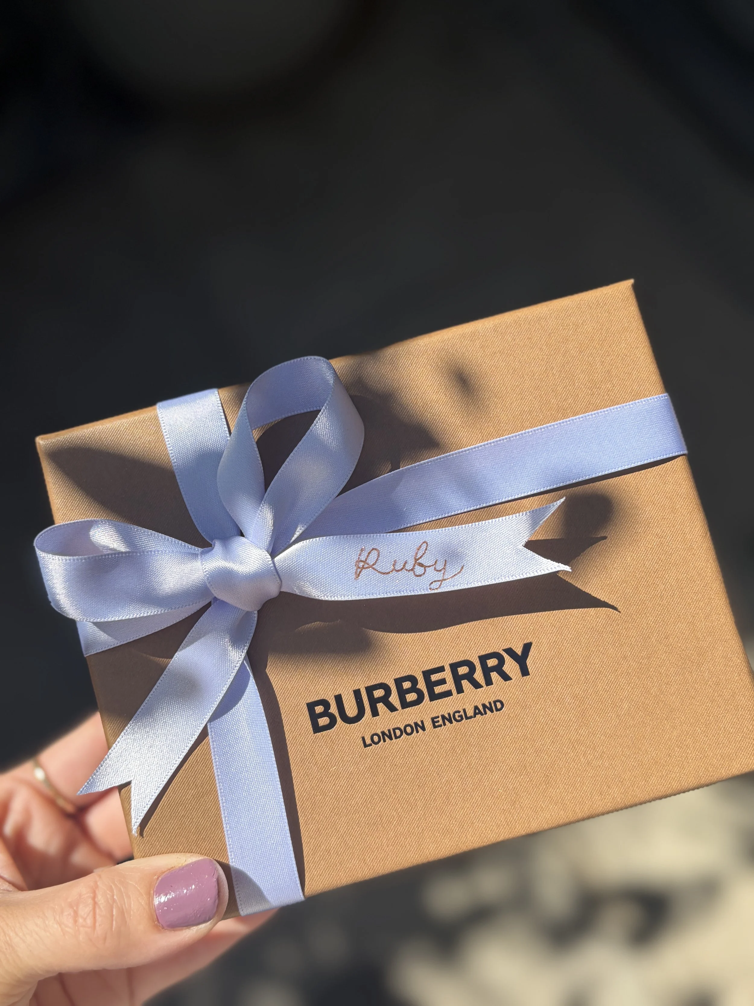 A hand holding a tan Burberry gift box with a lavender-coloured satin ribbon tied into a bow. The name 'Ruby' is written in gold foil in a cursive script on one end of the ribbon. The box has 'BURBERRY LONDON ENGLAND' written on it.
