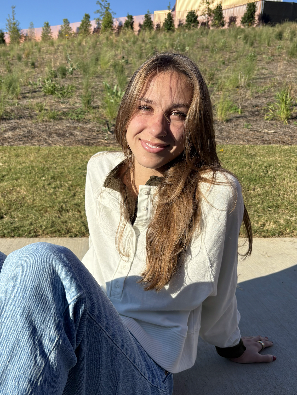 Young woman with long brown hair and a nose ring smiling, sitting on the sidewalk outdoors on a sunny day, with a grassy area and a fence with trees in the background.