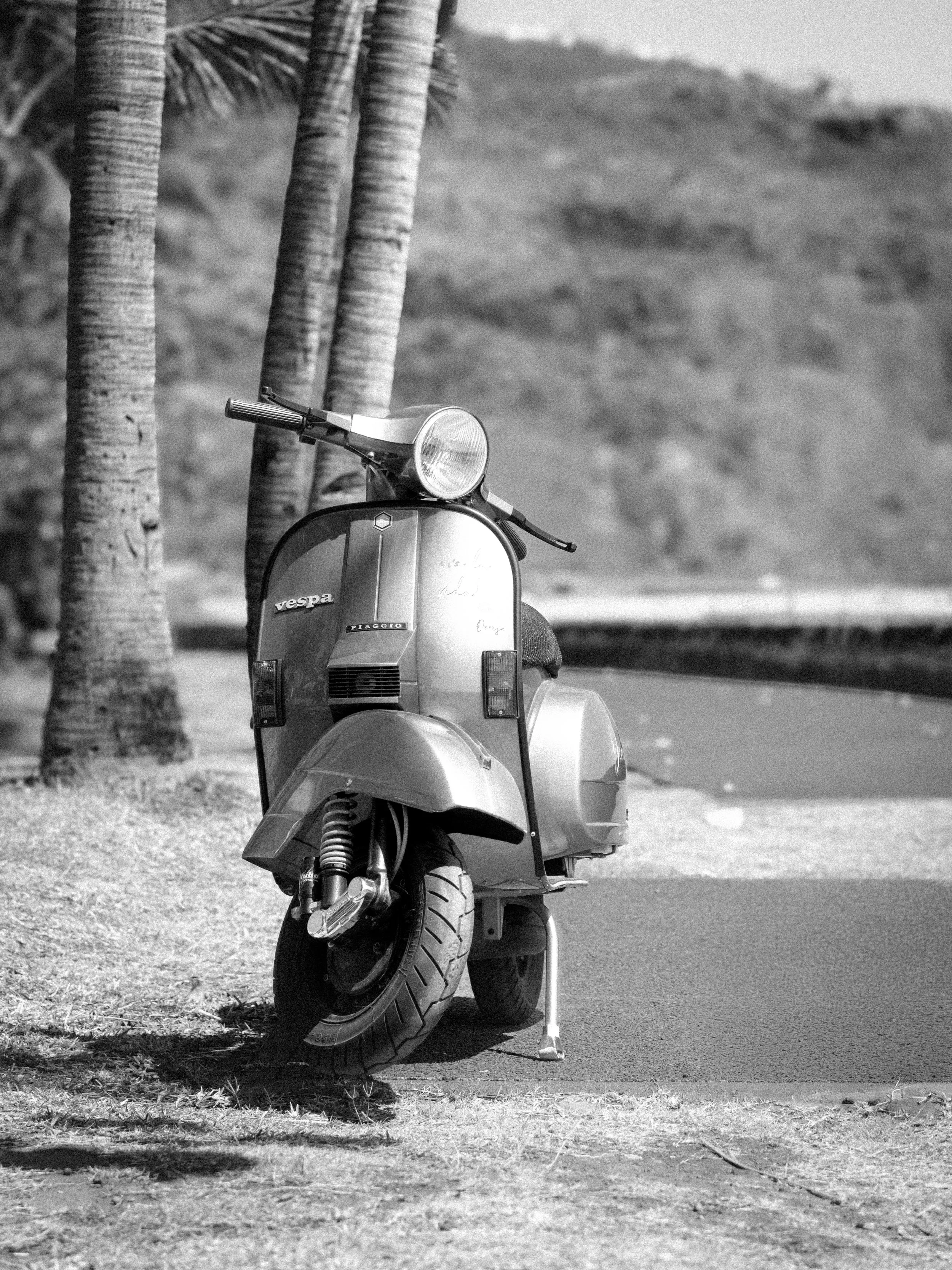 Scooter vintage stationné sur un trottoir, avec deux palmiers à proximité, en arrière-plan une colline ou montagne.