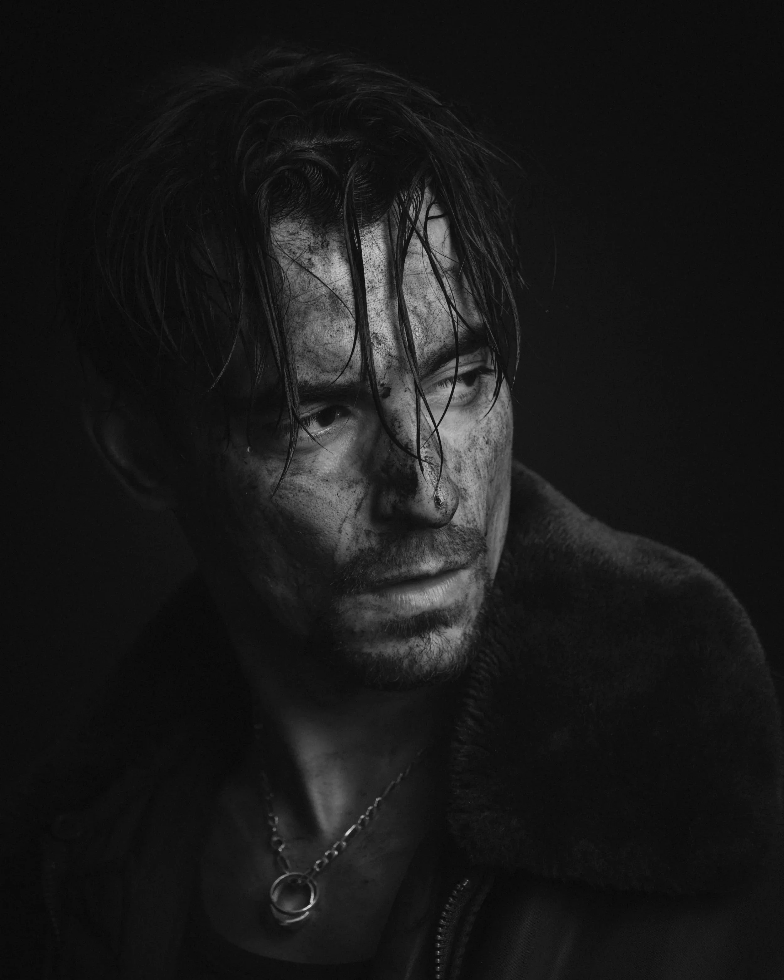 Un homme avec une expression intense, visage sale et cheveux en désordre, portant un collier en argent, en noir et blanc.