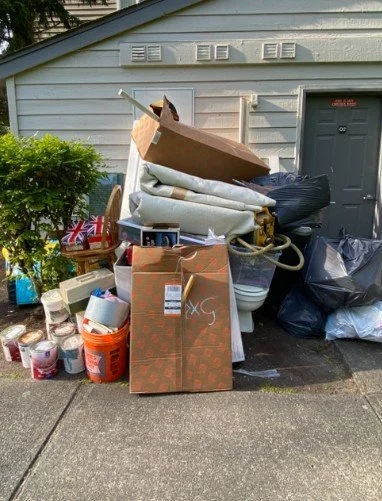 Overflowing pile of household items and trash near a garage door, including cardboard boxes, plastic bags, buckets, and spare household supplies.