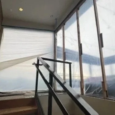 Interior of a building under construction with large windows, a staircase with a glass railing, and plastic sheeting covering part of the exterior.