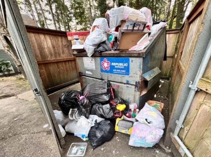 Overflowing dumpster filled with bags of trash, cardboard boxes, and debris outside a fenced area.