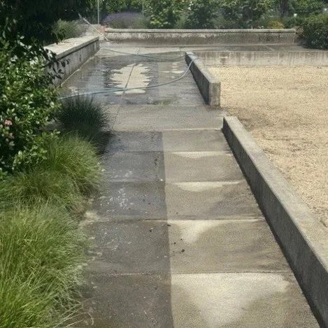 Sidewalk pressure washing service in progress with a narrow stream of water running alongside. Grass and bushes on the left, and a curb with gravel on the right.