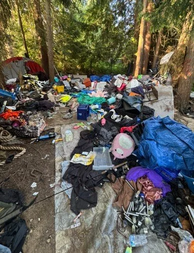 A cluttered outdoor scene with various camping or outdoor gear, clothes, and miscellaneous items scattered on the ground in a wooded area.