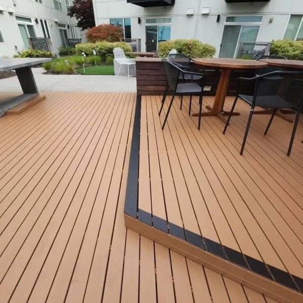 Outdoor wooden deck with black paint making the step visible, patio furniture including a round wooden table and black chairs, and a small garden area with bushes and trees in the background.