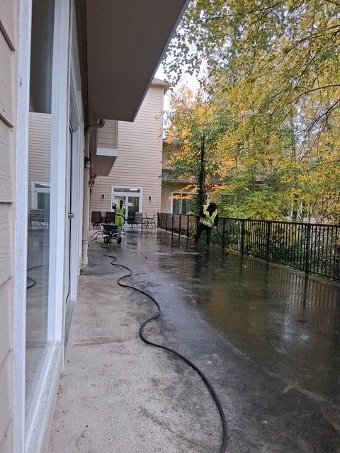Three workers in yellow safety vests cleaning an outdoor balcony or walkway with a hose on a rainy day. There are trees with fall foliage in the background.