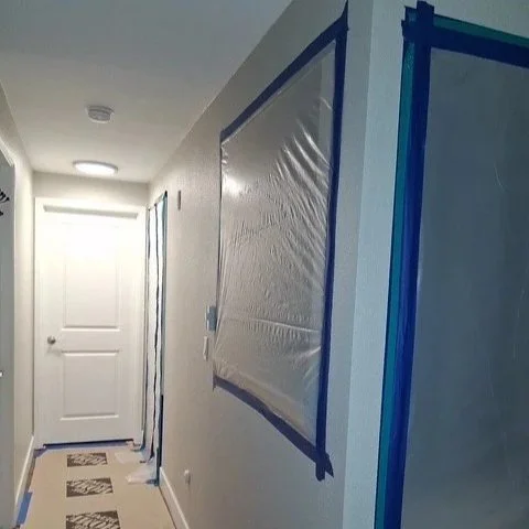 Interior hallway with a closed white door at the end, plastic-covered window, and protective plastic on the walls, suggesting ongoing renovation or painting.