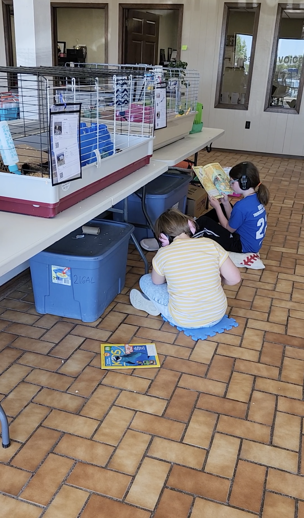 Two children sitting on the floor under a table reading books, with hamster cages and supplies on the table above them.