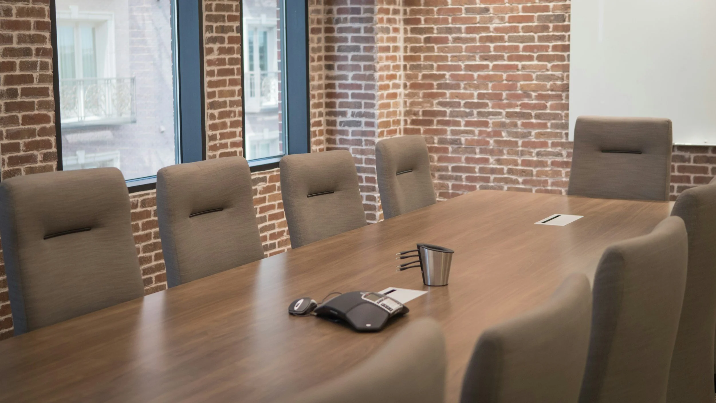 Empty conference room with brick walls, large windows, beige chairs around a wooden table, and office supplies on the table.