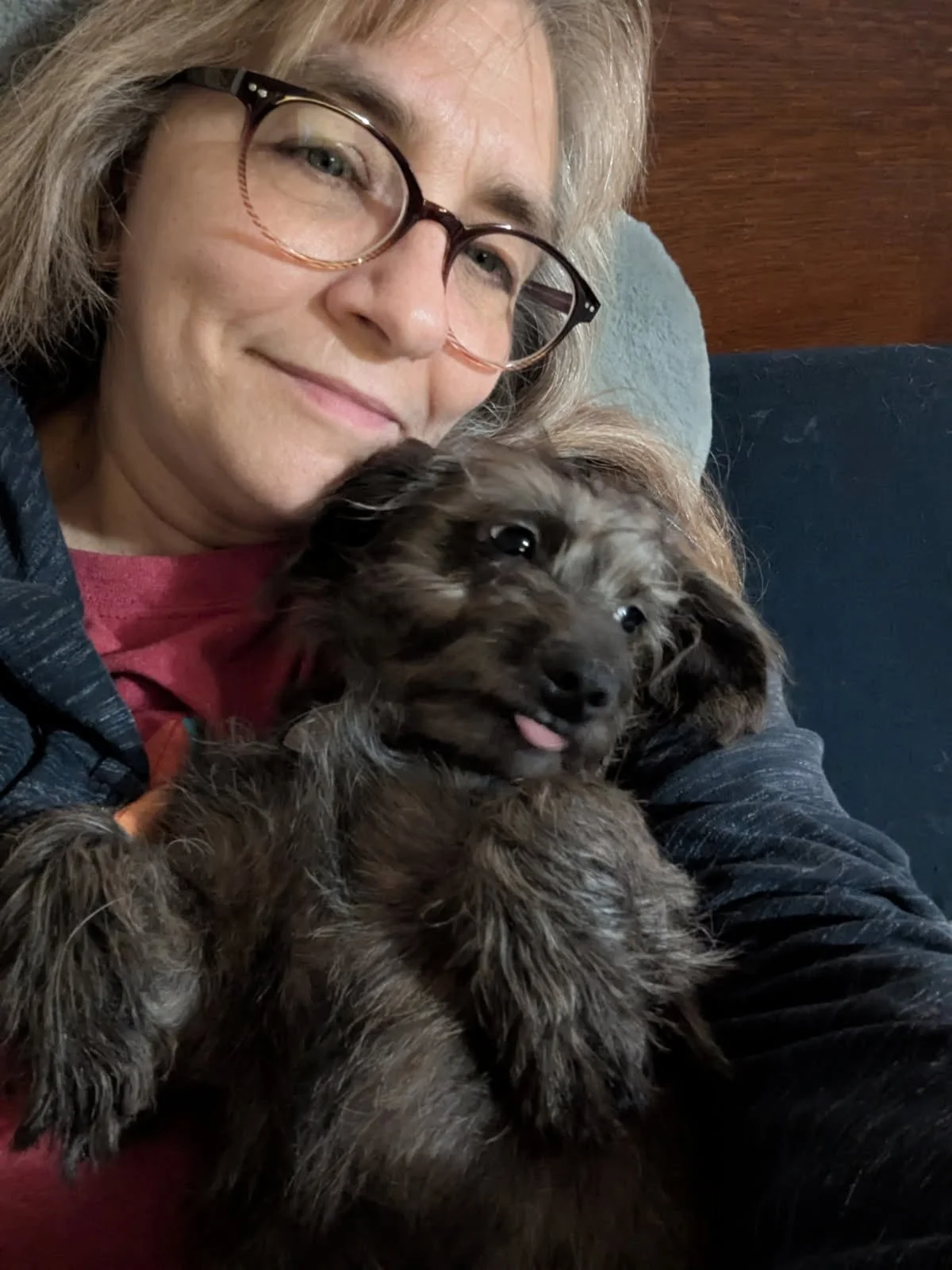 A woman with glasses and gray hair holding a small brown and black puppy with floppy ears and a tongue sticking out, sitting on a couch with a wooden background.