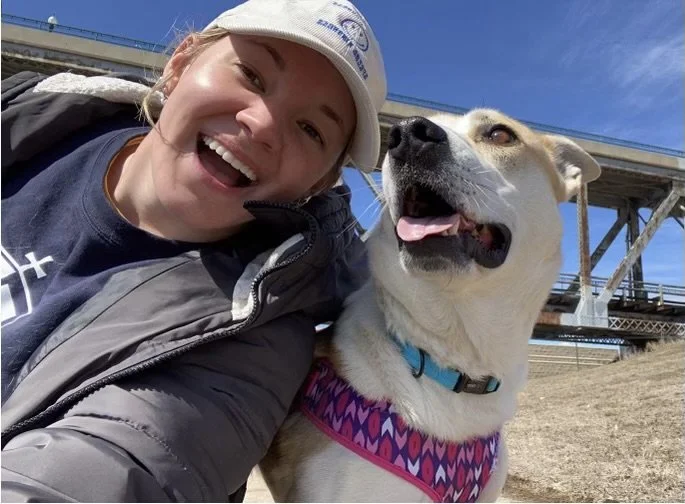 A woman smiling with a white dog wearing a colorful harness outdoors under a bridge.