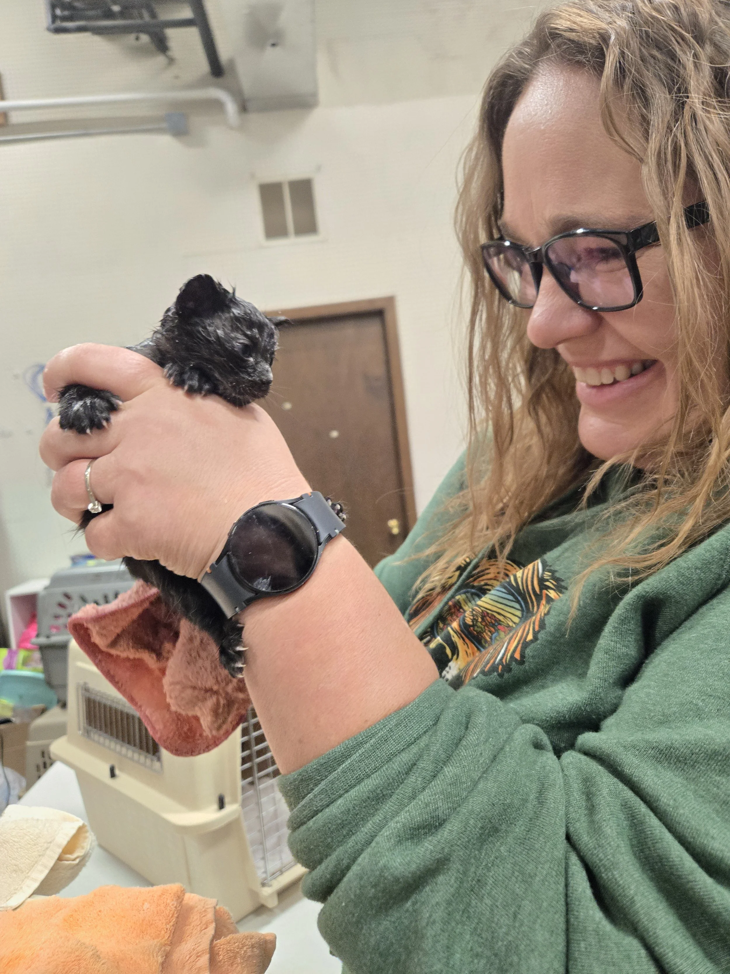 A woman with glasses smiling while holding a small black puppy in her hands. The woman is wearing a green shirt and a black watch, and is in an indoor setting with pet cages and supplies in the background.