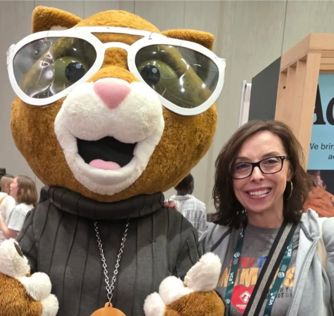 A woman smiling and standing next to a large anthropomorphic animal mascot with glasses, a pink nose, and a big open mouth, at a convention or event.