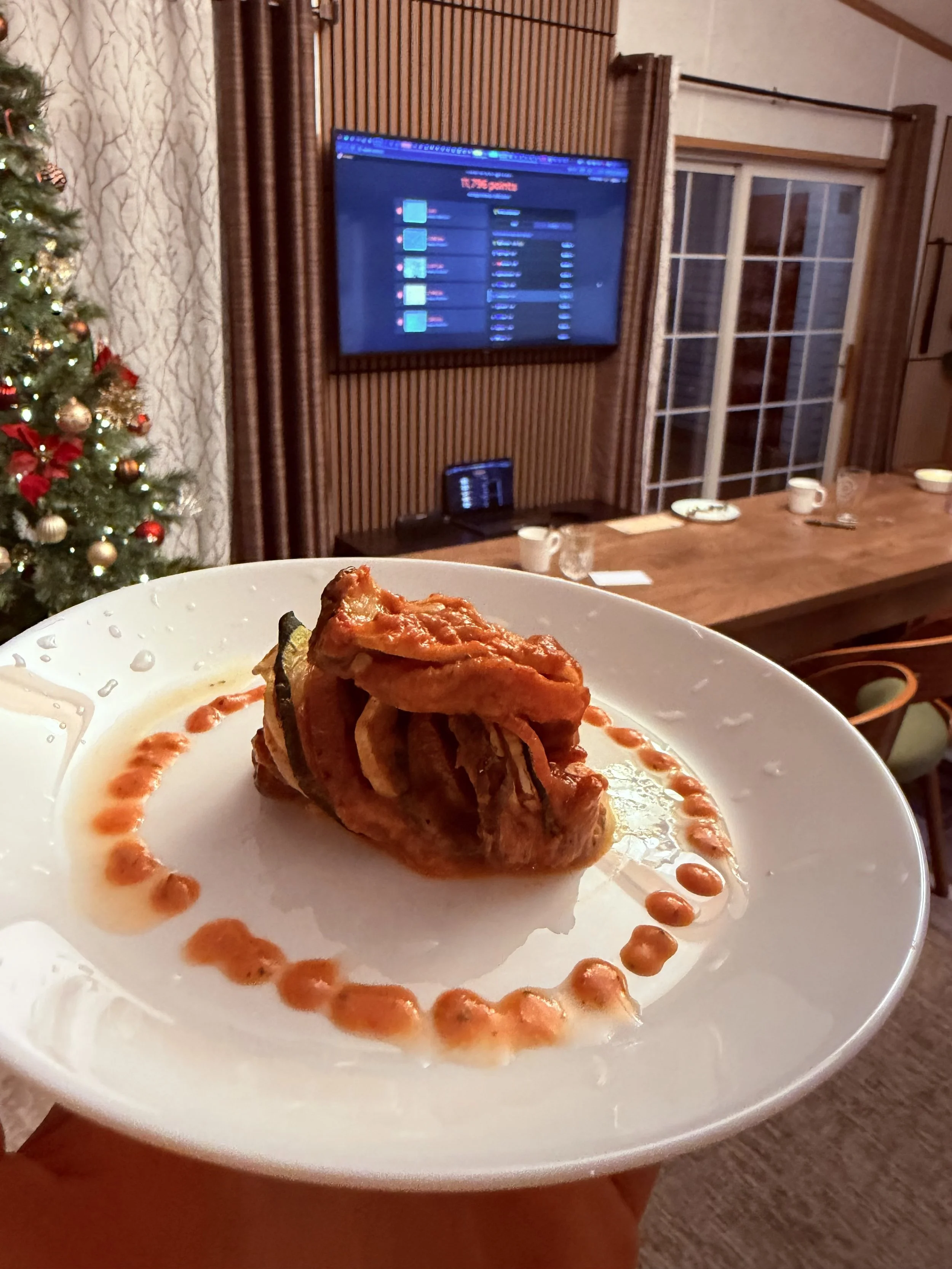 A plate of food featuring a fried chicken drumstick with a red sauce drizzle and a decorative red sauce border on the plate. In the background, there is a living room with a decorated Christmas tree, a flat-screen TV mounted on the wall, a wooden table with cups and dishes, a sliding glass door, and curtains.